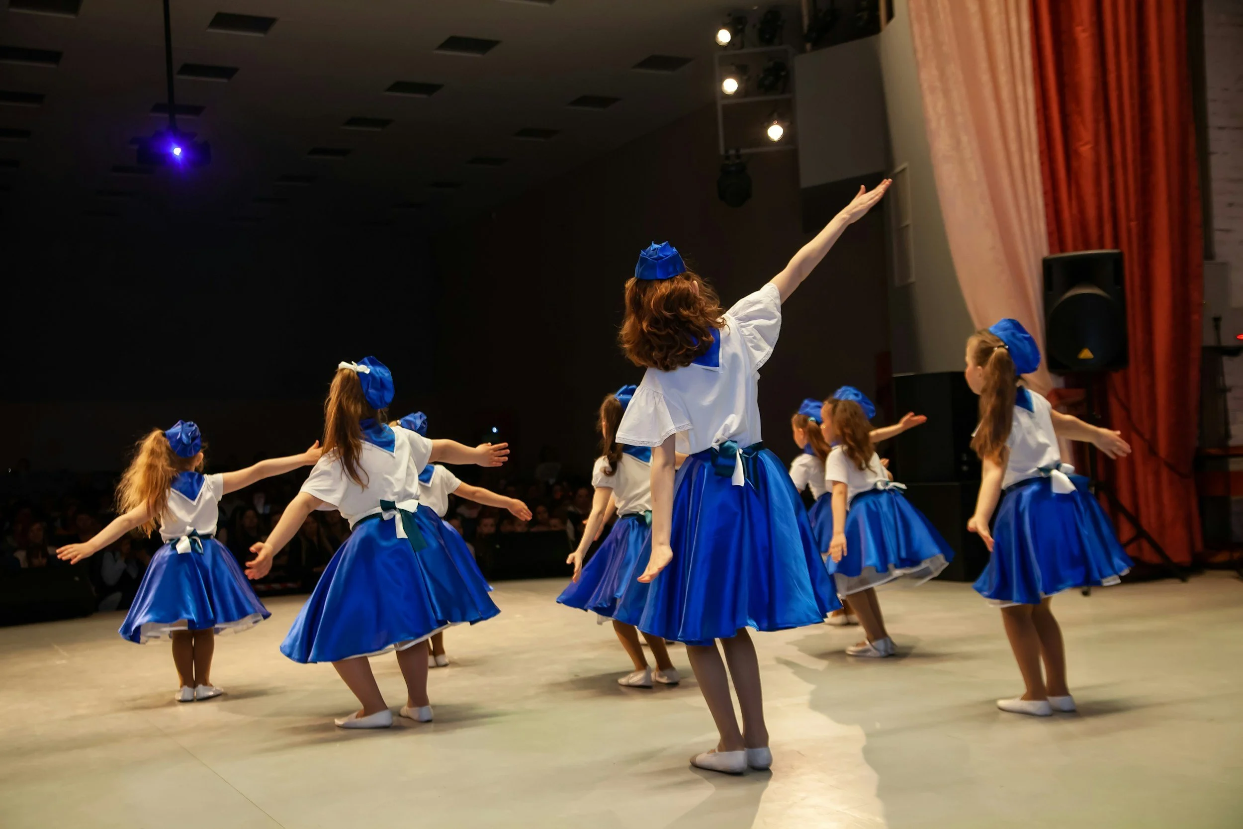 Group of young girls performing a dance routine on stage, wearing blue skirts, white blouses, and matching blue headbands, with one girl leading the dance with her arm raised.
