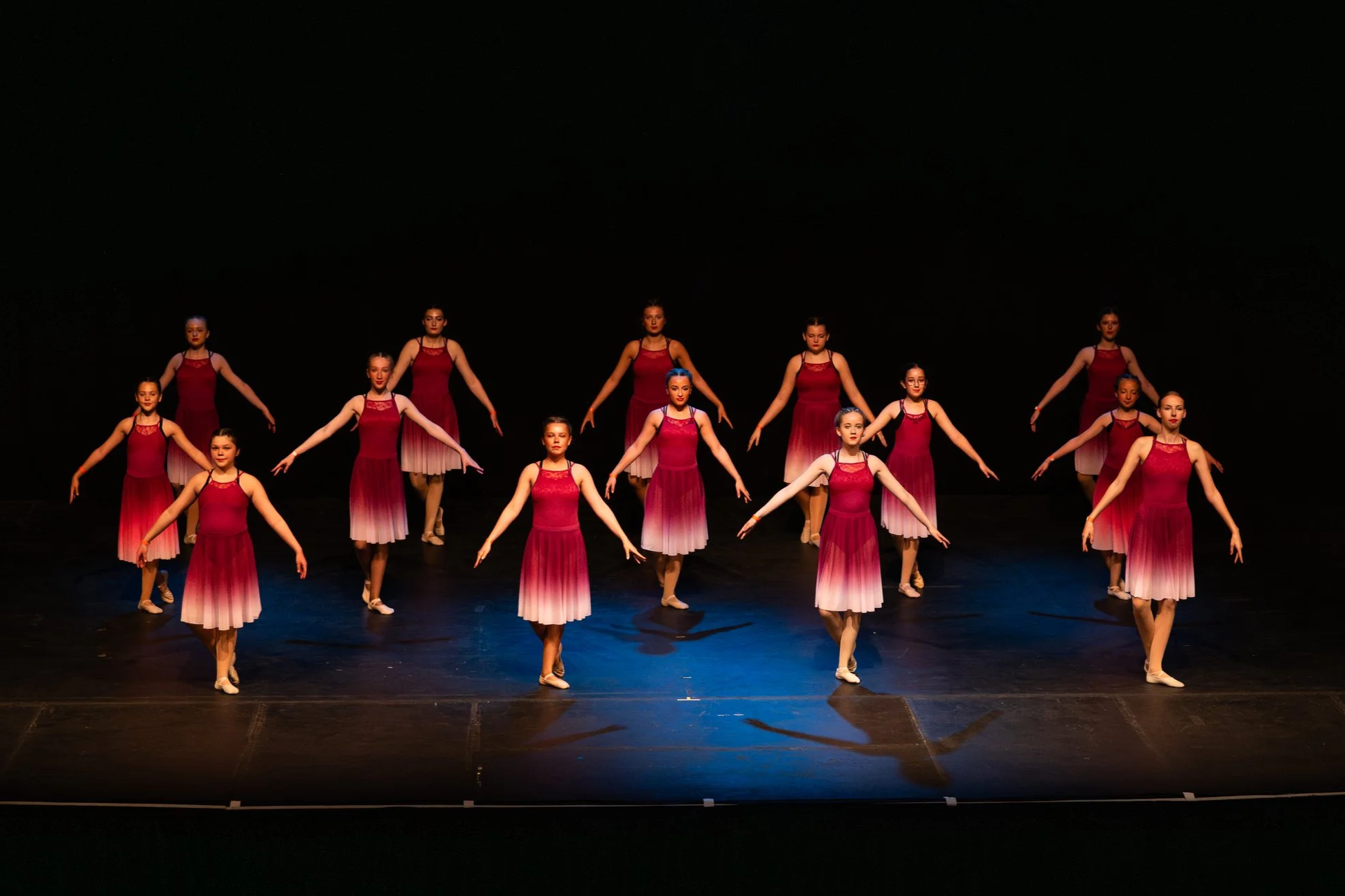 Children dressed in white tutus and tops performing a dance on stage with colorful lighting, audience in background.