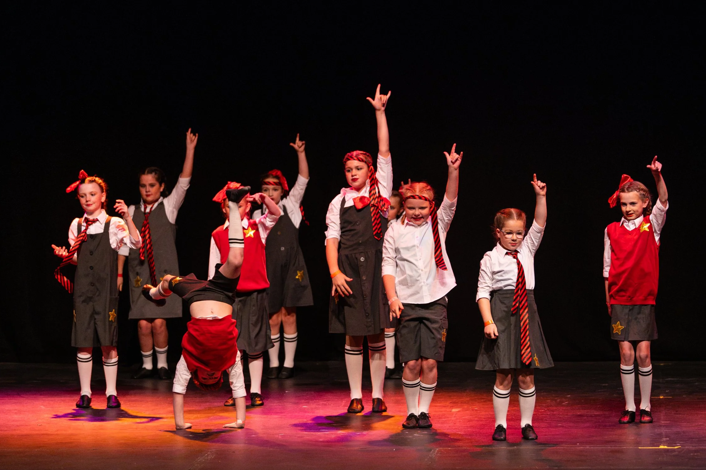 Group of young girls performing a dance routine on stage, wearing blue skirts, white blouses, and matching blue headbands, with one girl leading the dance with her arm raised.