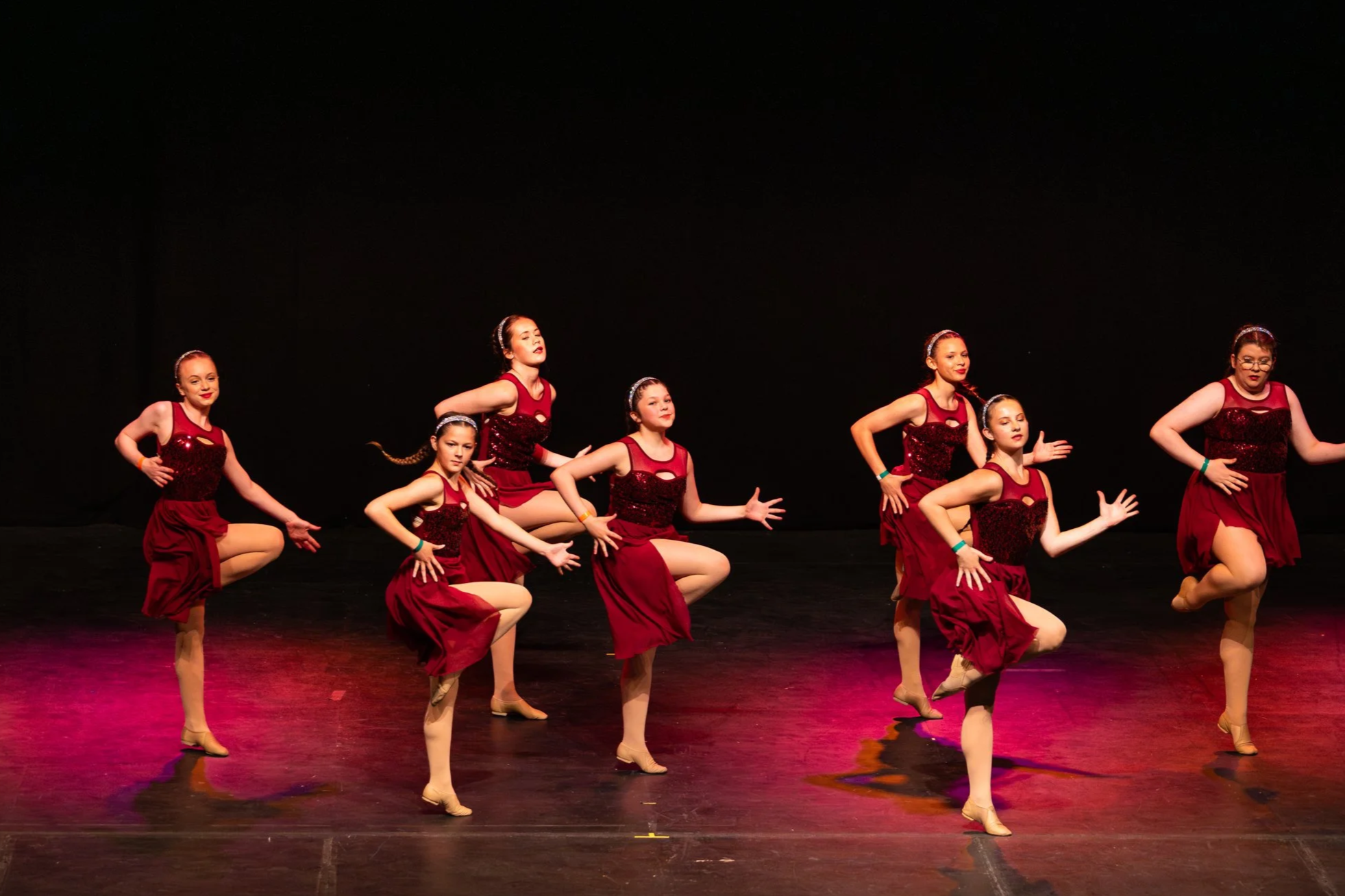 A group of dancers performing on stage in black costumes with a dark background. Light patterns are projected on the wall behind them.