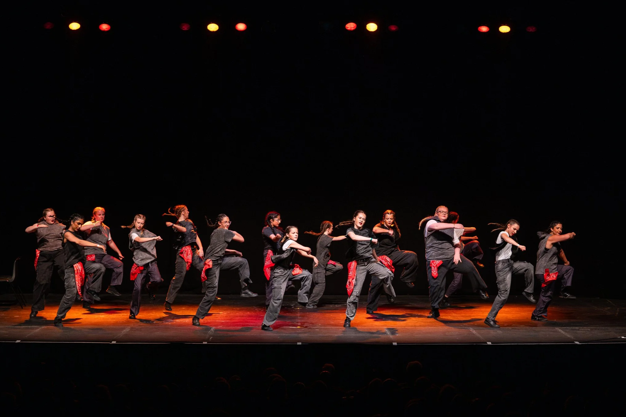 A group of seven male dancers on stage performing a choreographed dance routine, illuminated by colorful stage lights with a dark background.