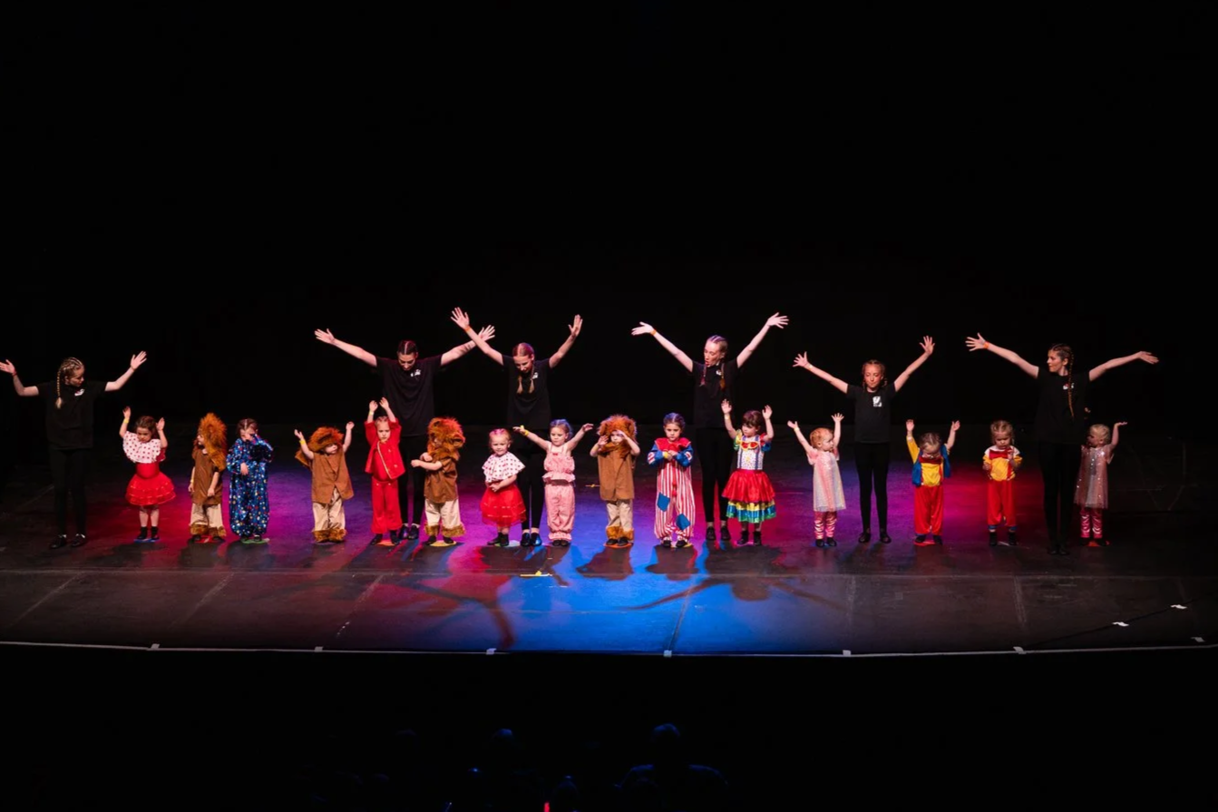 Young girls in colorful tutus and yellow T-shirts performing a dance on stage with curtains and a screen in the background.