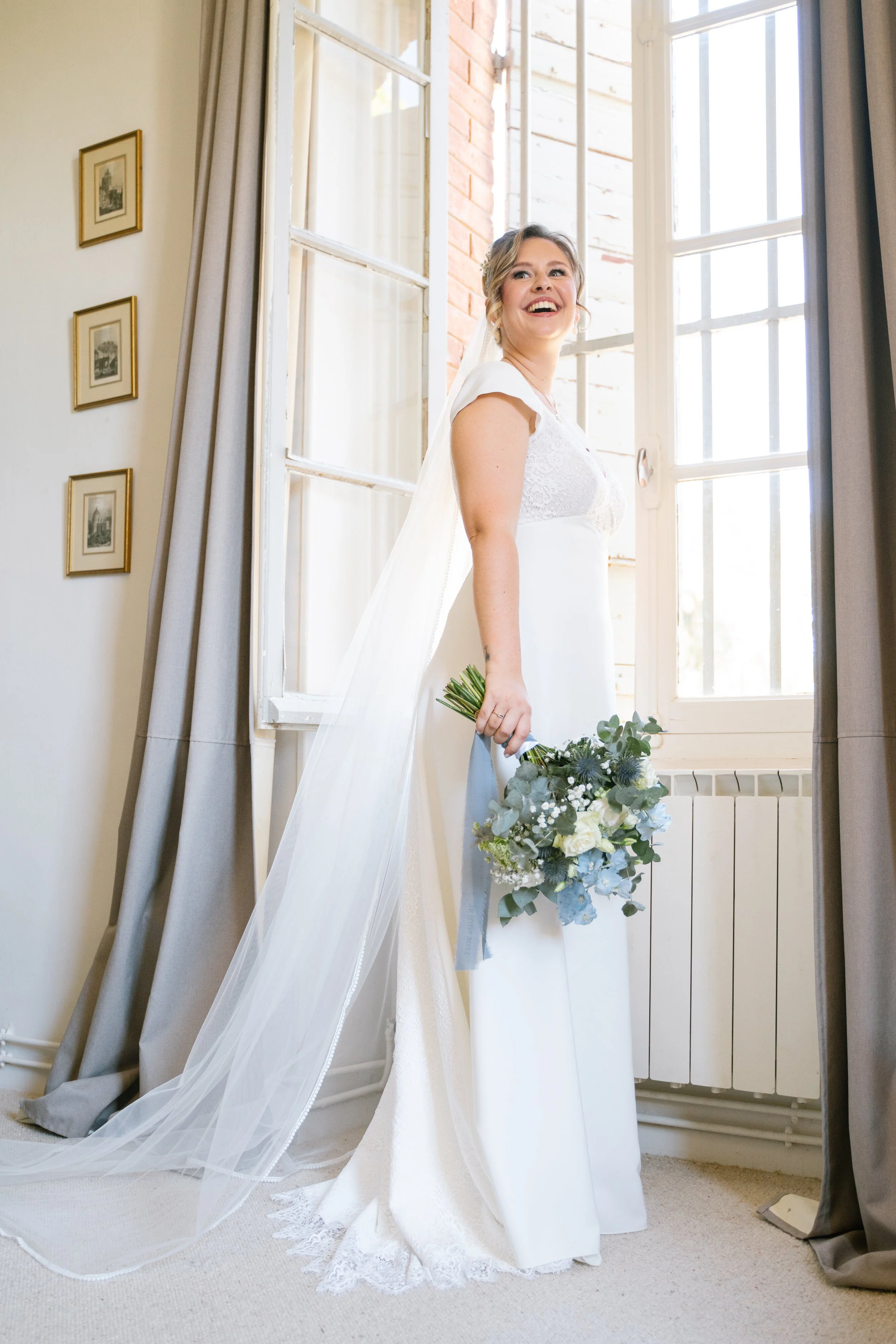 A bride in a white wedding dress holding a bouquet, standing next to a window, smiling.