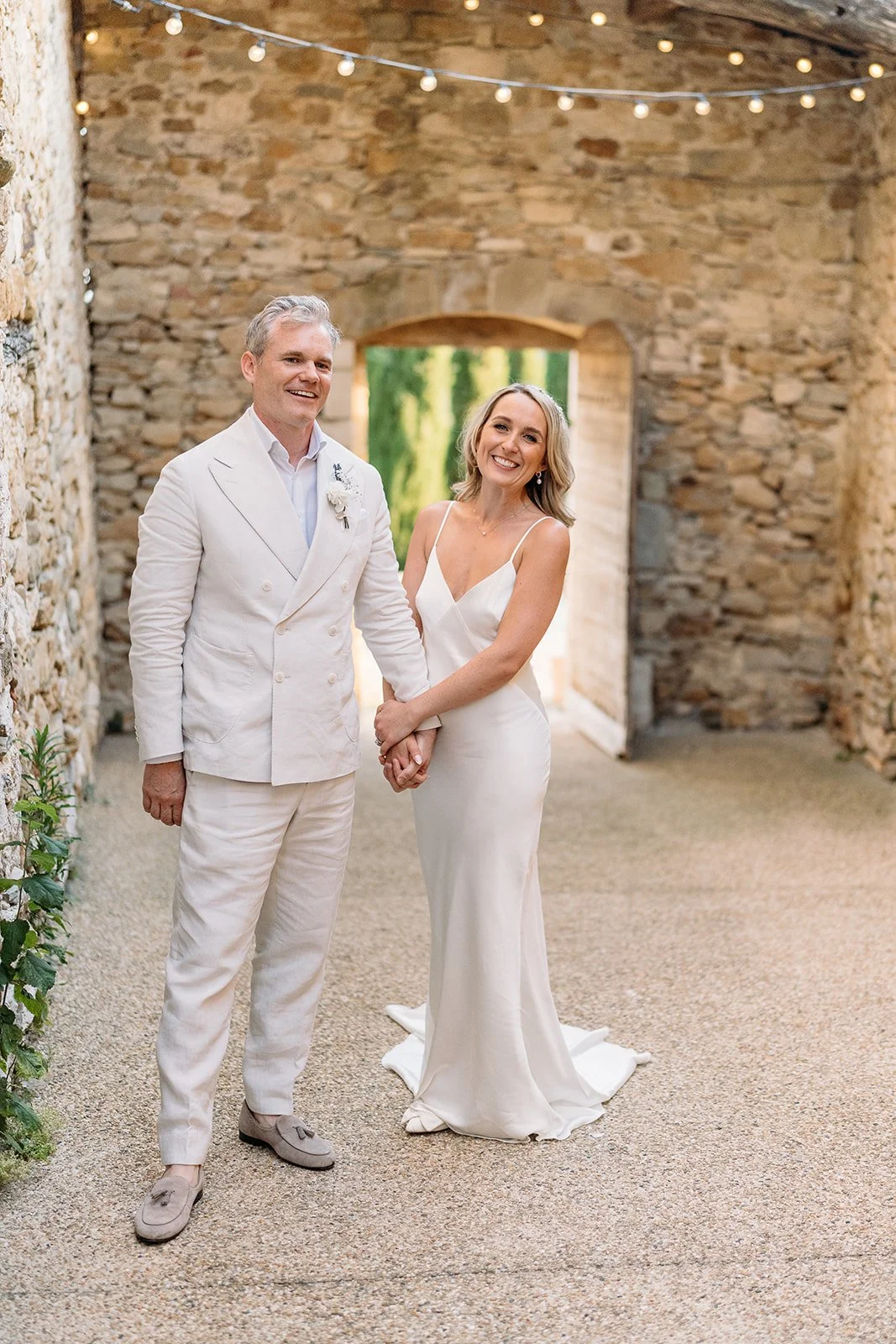 Bride and groom holding hands in a stone courtyard at Le Clos du Tuillier, long-lasting natural wedding makeup by Mathilde Vintze