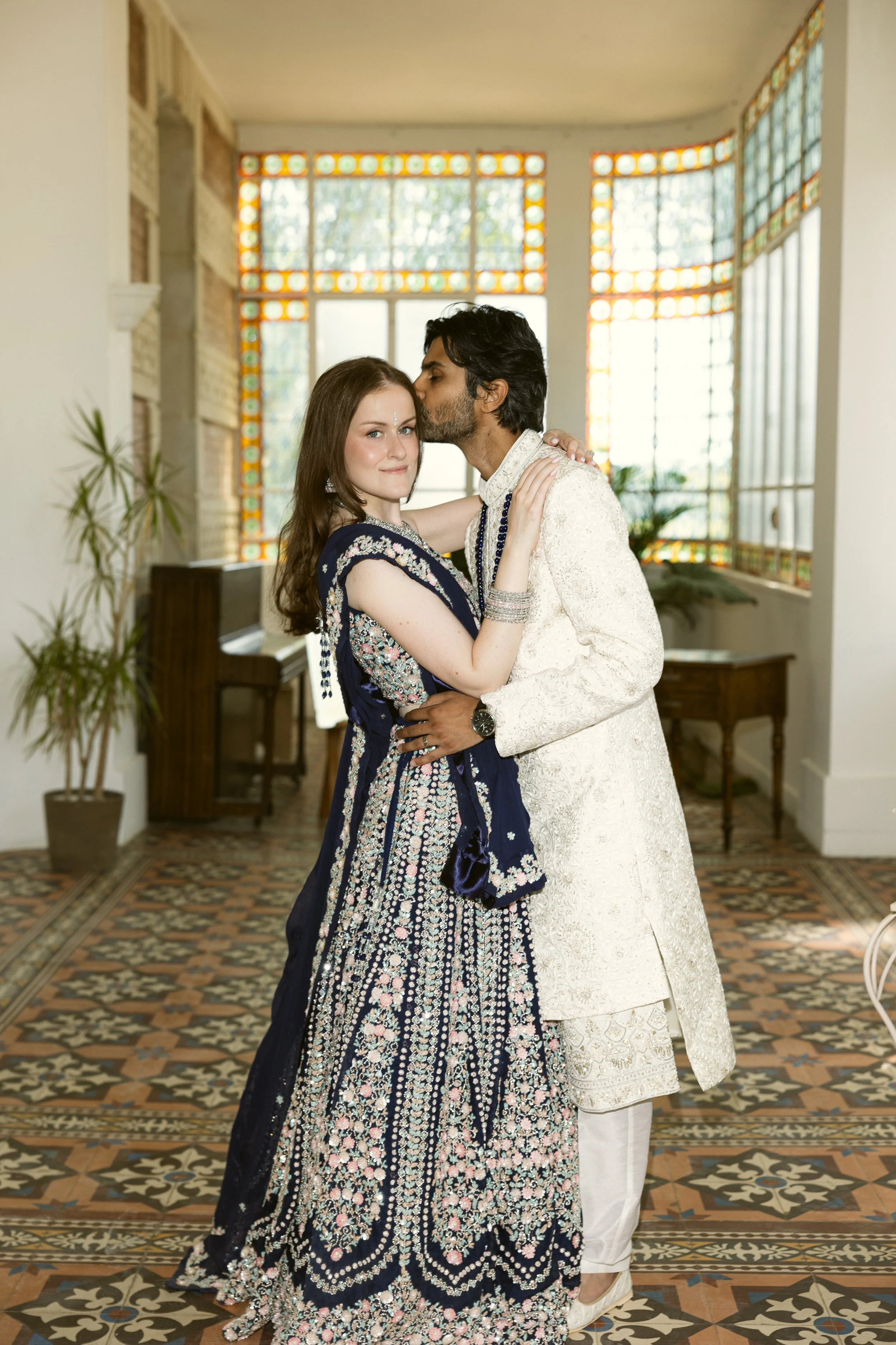 Indian wedding portrait of the bride in a hand-embroidered navy lehenga and the groom in an ivory sherwani, framed by stained glass and patterned tiles. Refined bridal makeup by Mathilde Vintze Artistry.