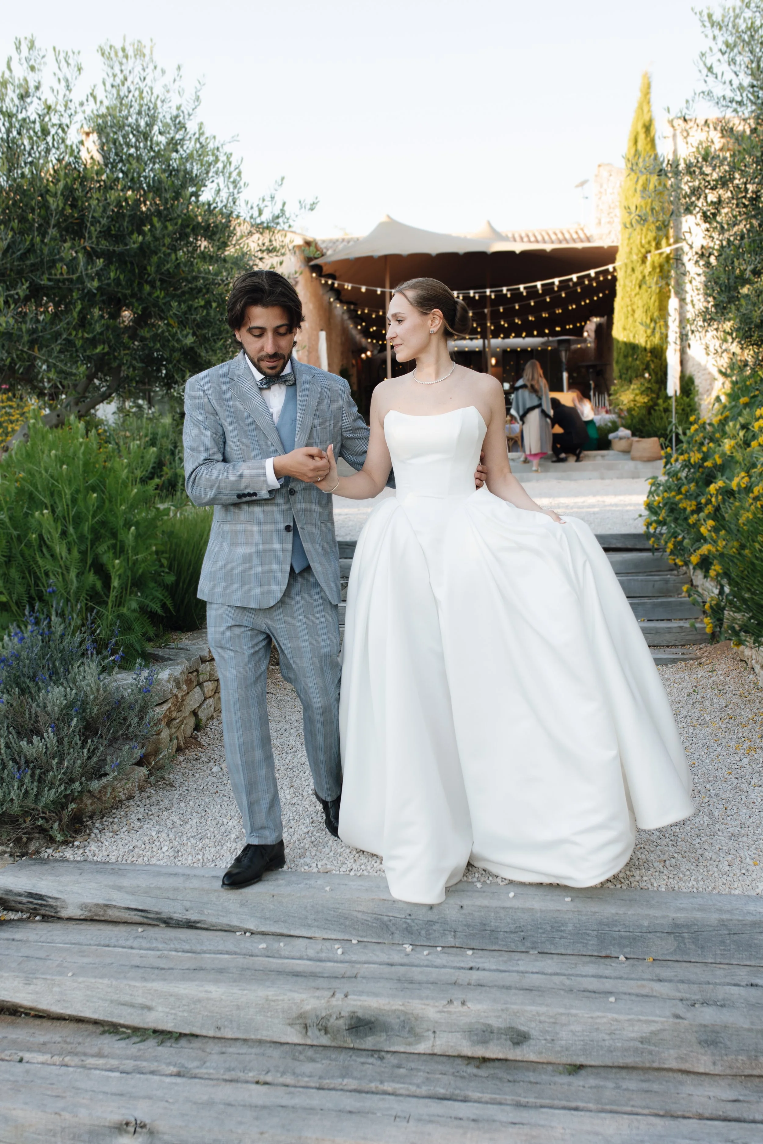 Elegant bride and groom portrait in a sunlit garden, with the bride wearing radiant long-lasting wedding makeup by Mathilde Vintze Artistry alongside her groom in a grey suit.