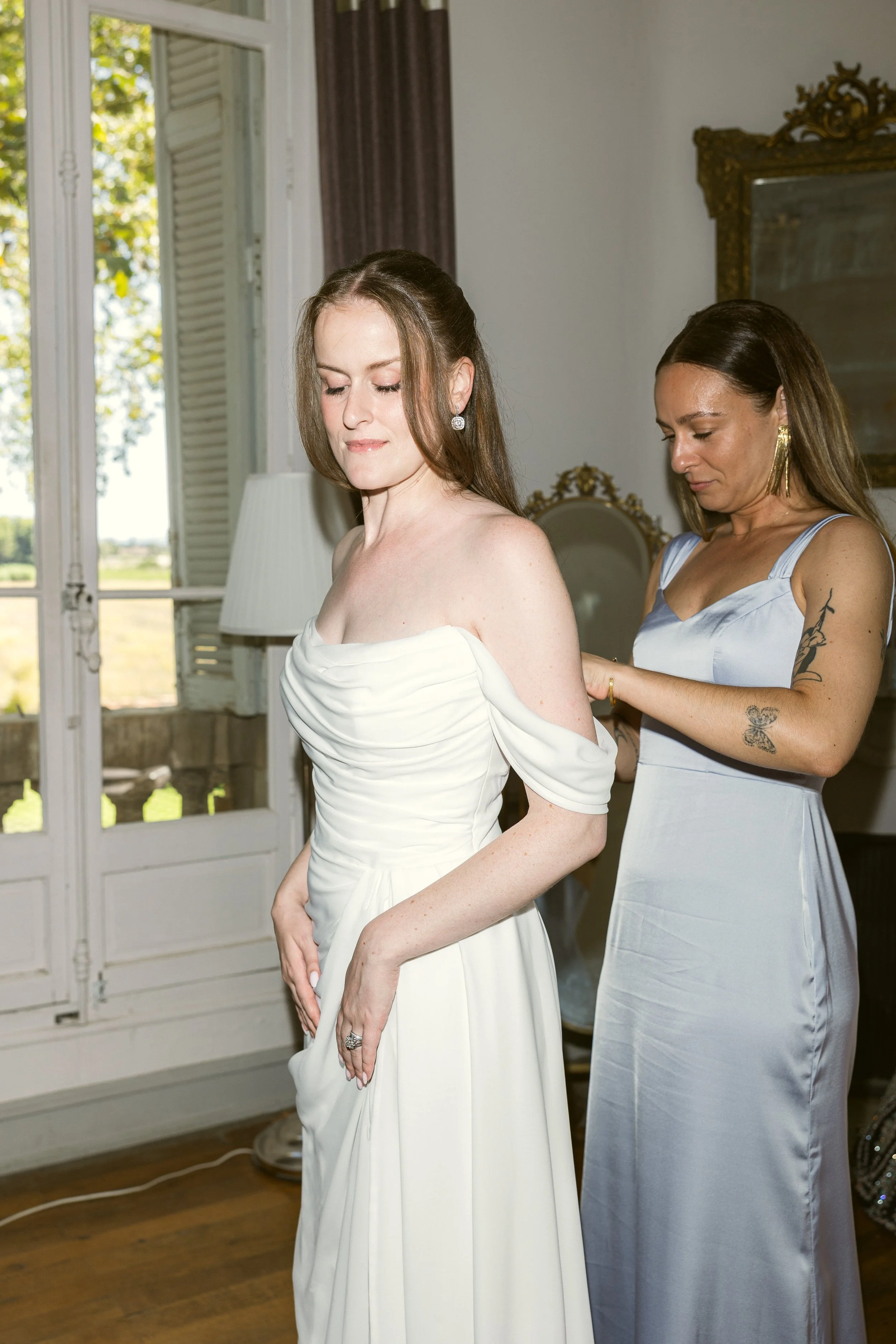 Final touches before the ceremony: a bridesmaid fastens the bride's off-the-shoulder wedding gown. Soft, radiant bridal makeup that reveals rather than covers.