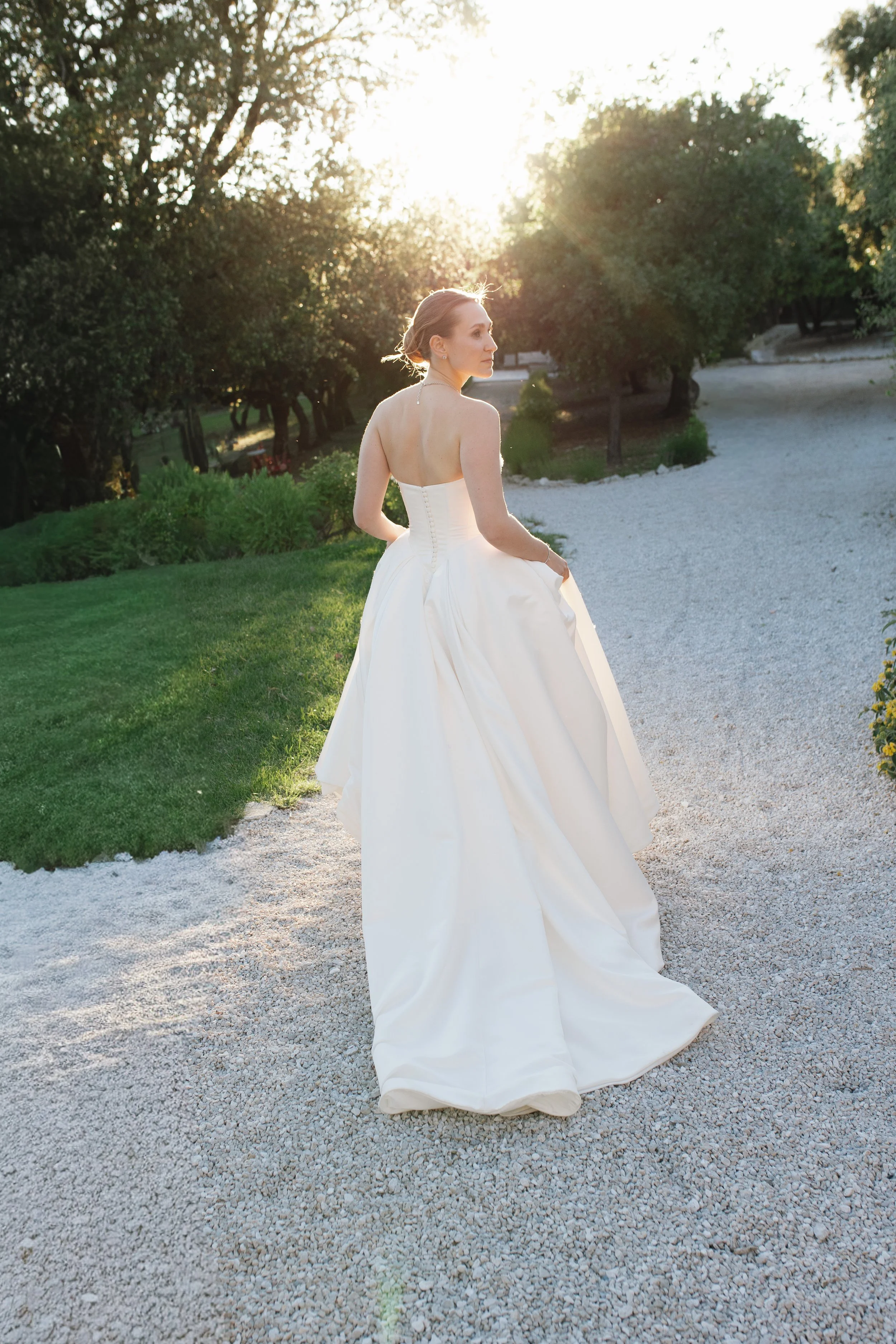 Bride from behind in a flowing open-back gown during golden hour, her sleek bridal hairstyle and natural makeup by Mathilde Vintze Artistry on full display.