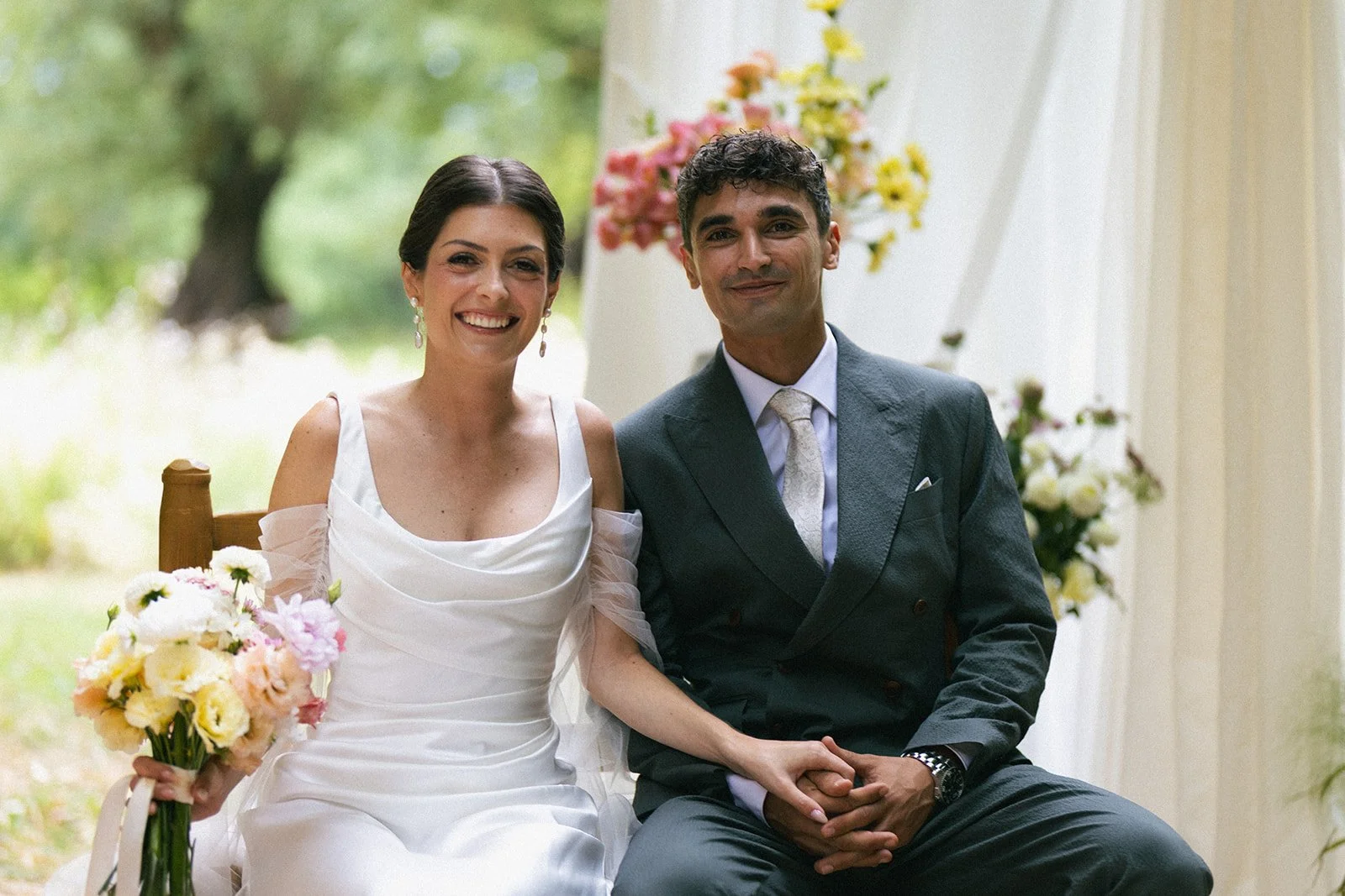 Bride and groom seated side by side, smiling in front of summer flowers at their wedding in Provence. Fresh, glowing bridal makeup made for outdoor summer light.
