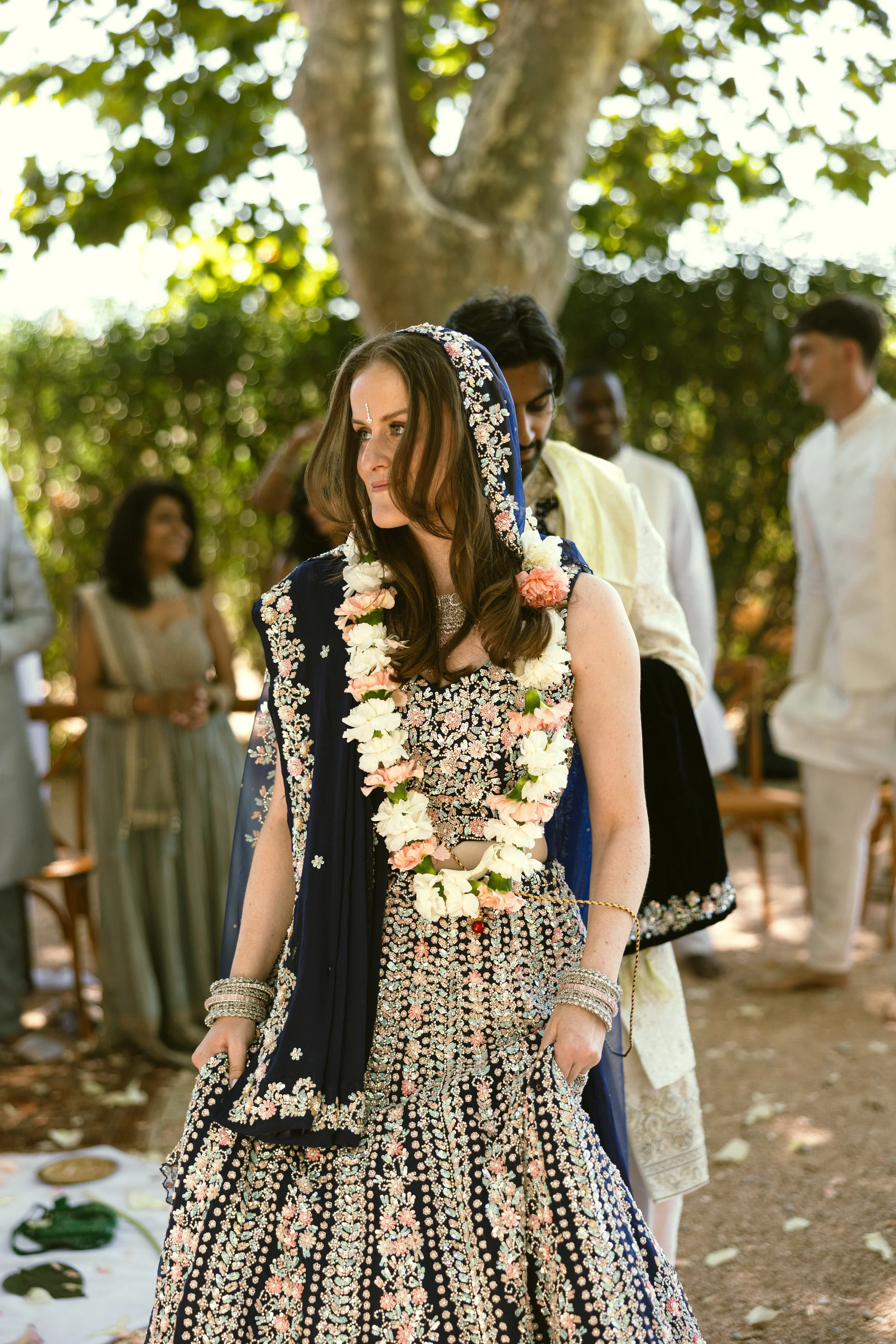 The bride during her outdoor Indian wedding ceremony, wearing a fresh flower garland and an embroidered navy lehenga with a draped dupatta veil. Soft, long-wearing bridal makeup by Mathilde Vintze Artistry, bridal makeup artist in Provence.