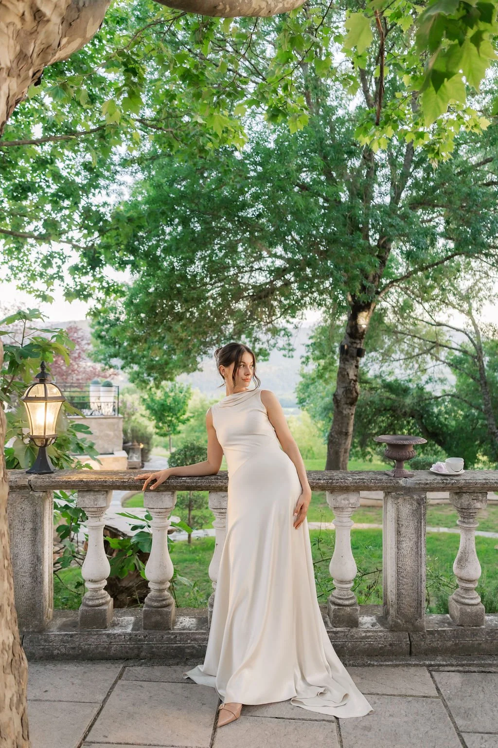 Bride alone on a stone terrace under the trees in Provence, flowing gown and elegant natural wedding makeup by Mathilde Vintze