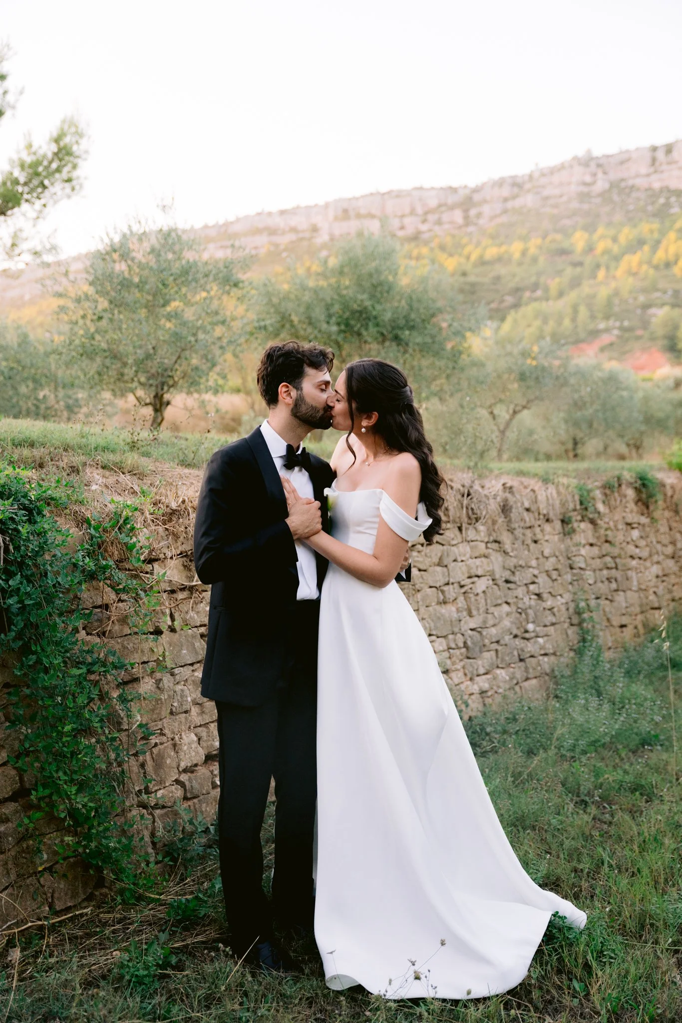 A bride and groom kissing outdoors with a stone wall and greenery in the background.