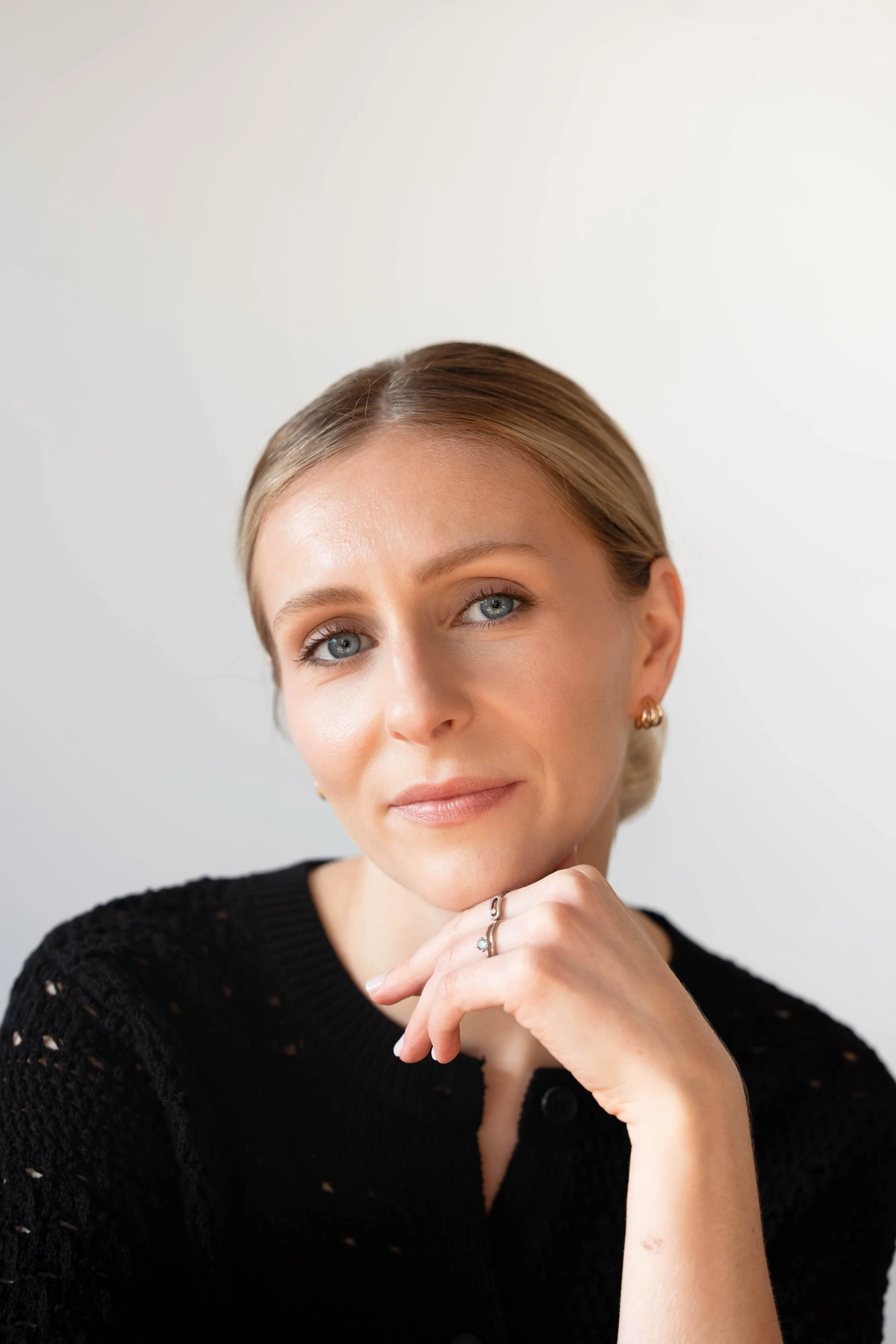 A woman with blonde hair and blue eyes, wearing a black knit top and gold earrings, poses with her hand resting under her chin against a plain white background.