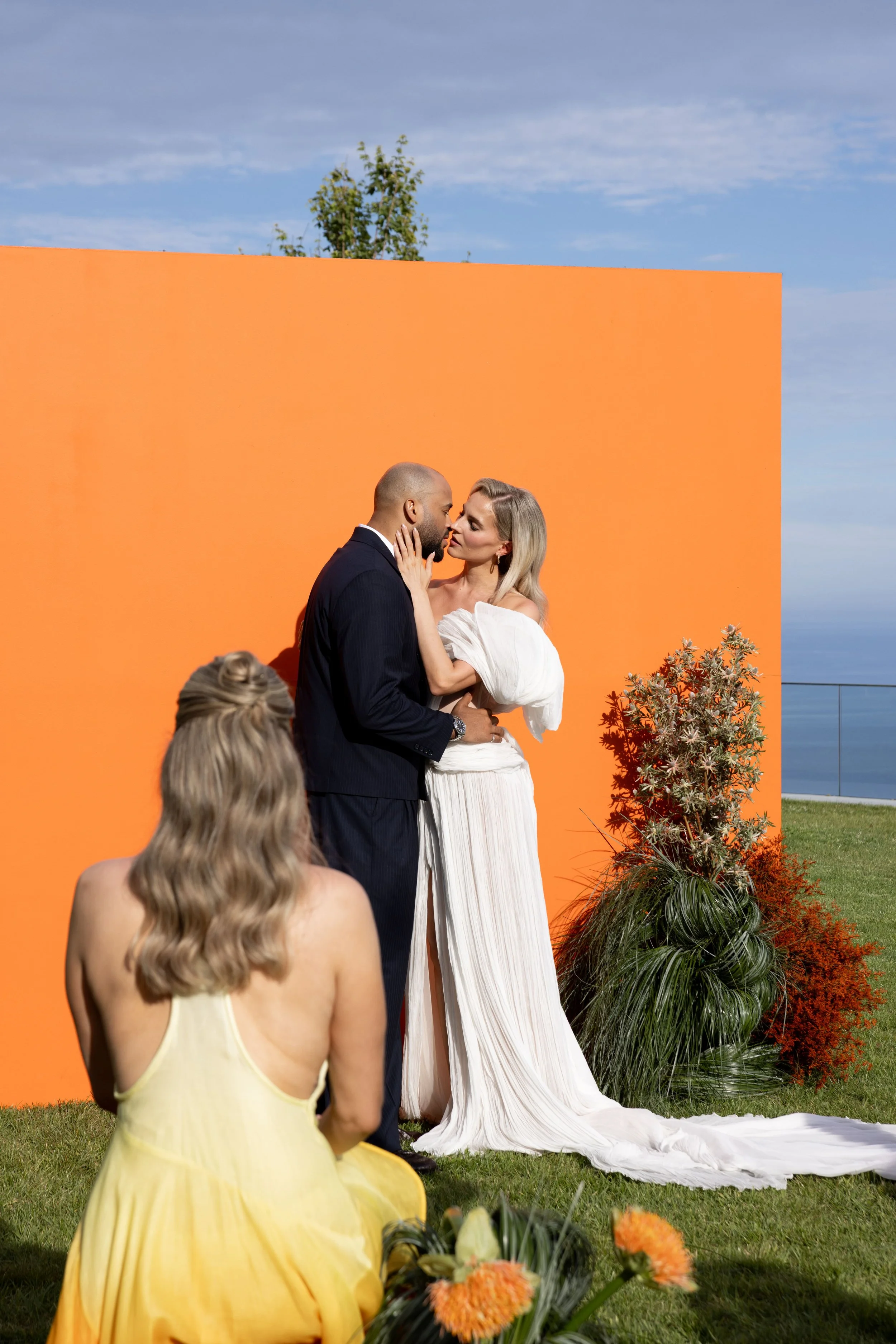 Bride and groom embracing at the orange ceremony backdrop seen past a guest in yellow, Côte d'Azur wedding makeup by Mathilde Vintze