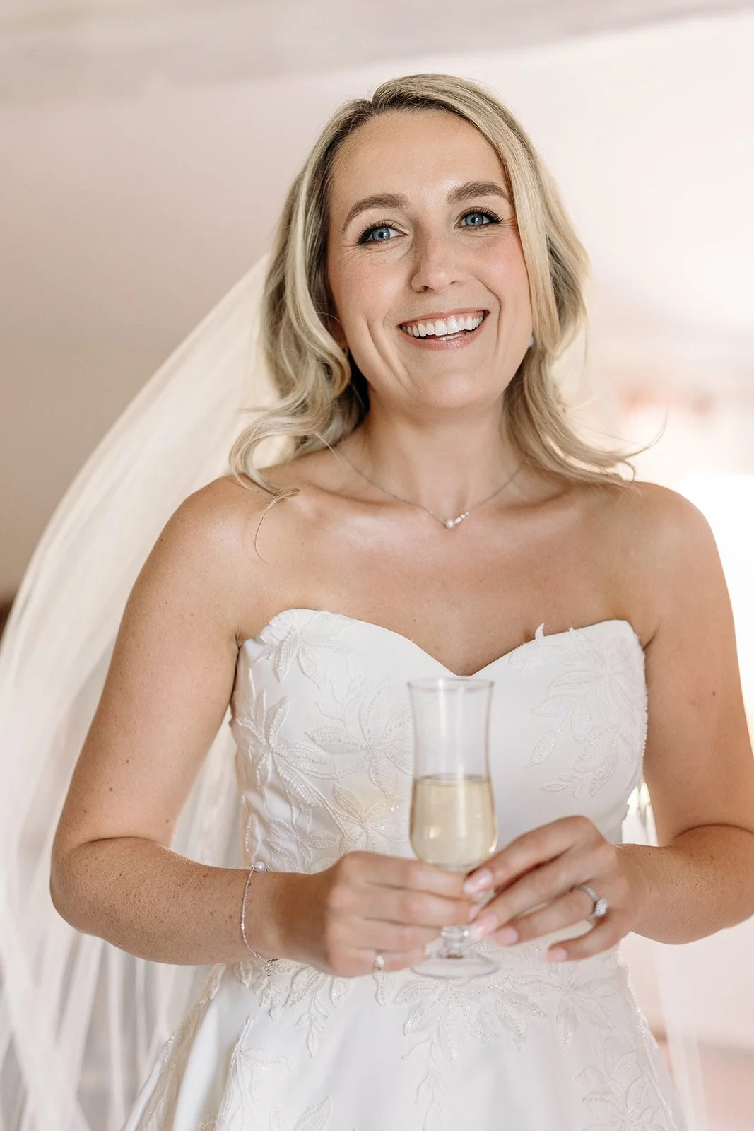 Bride in an embroidered gown and veil holding a glass of champagne, radiant natural bridal makeup in Provence by Mathilde Vintze