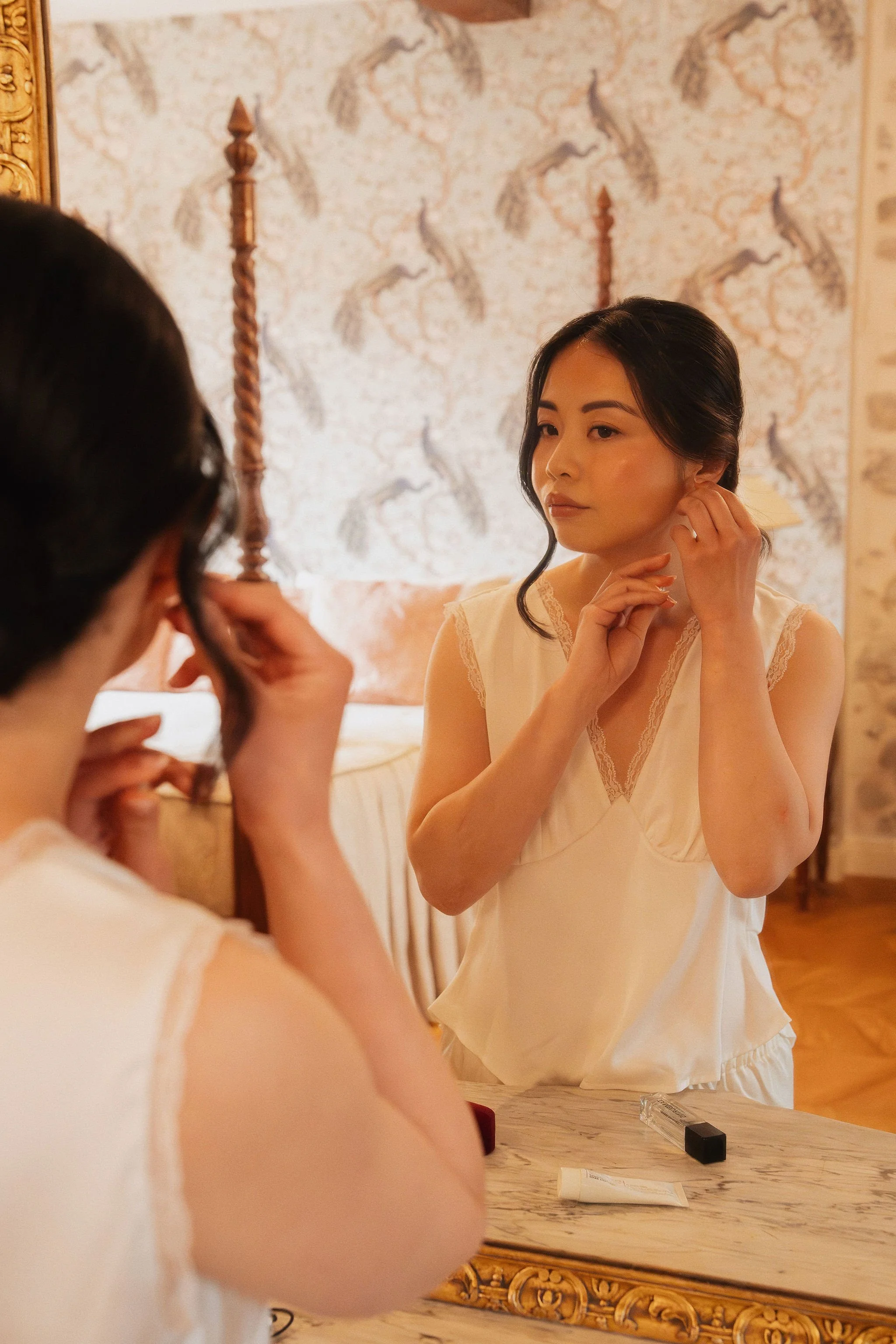 Bride putting on earrings in the mirror while getting ready, elegant bridal makeup by Mathilde Vintze