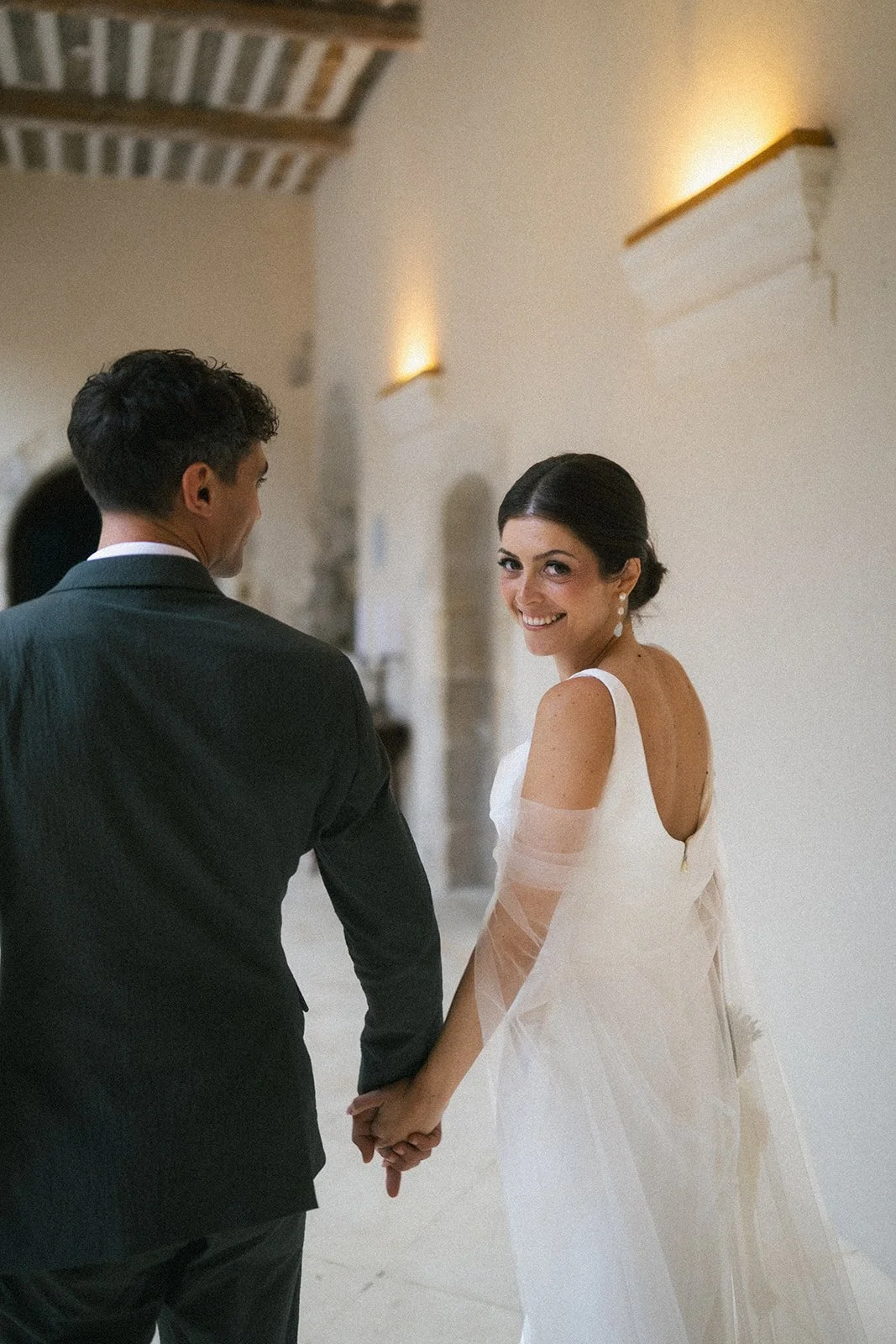 Bride and groom walking hand in hand through the vaulted halls of Couvent Notre-Dame des Prés, the bride glancing back over her shoulder in a low-back wedding dress.