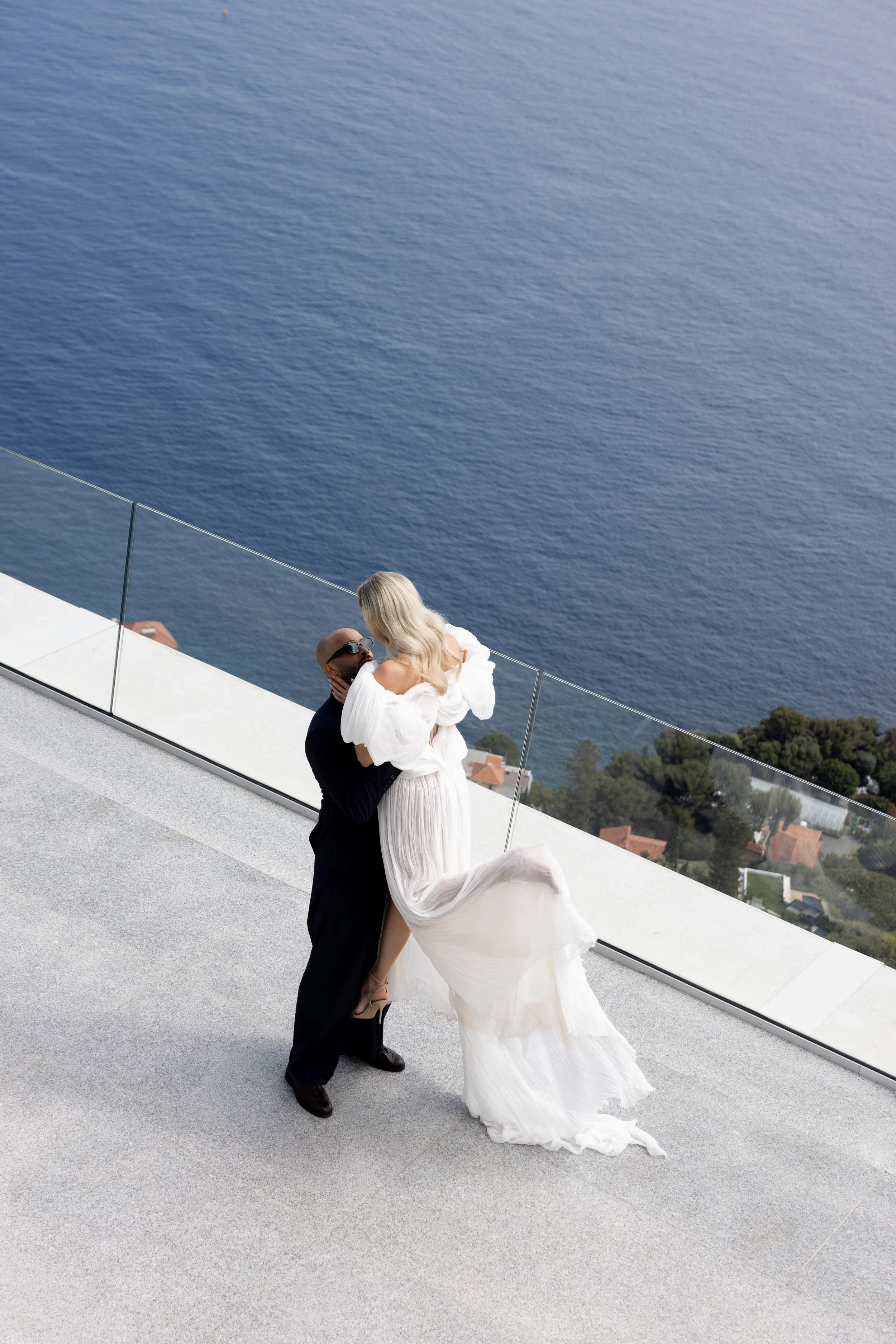 Bride and groom on the terrace high above the Mediterranean, the bride's dress in the breeze, French Riviera bridal makeup by Mathilde Vintze