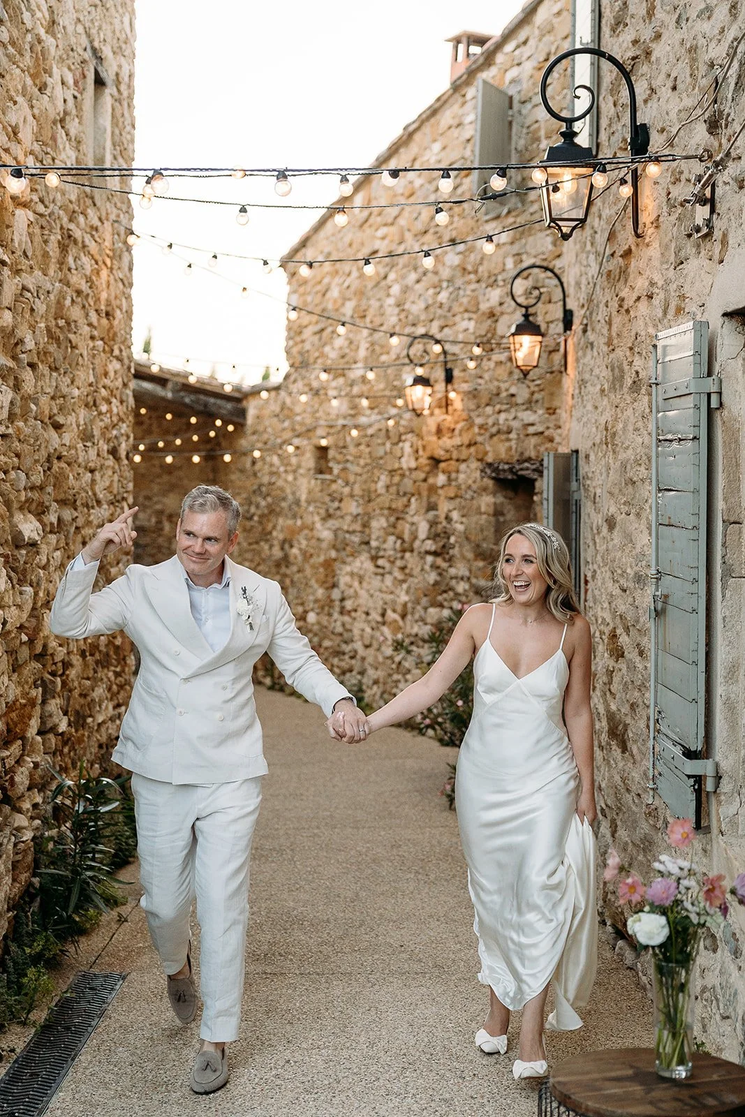 Bride and groom hand in hand under festoon lights in a stone lane at Le Clos du Tuillier, natural wedding makeup by Mathilde Vintze
