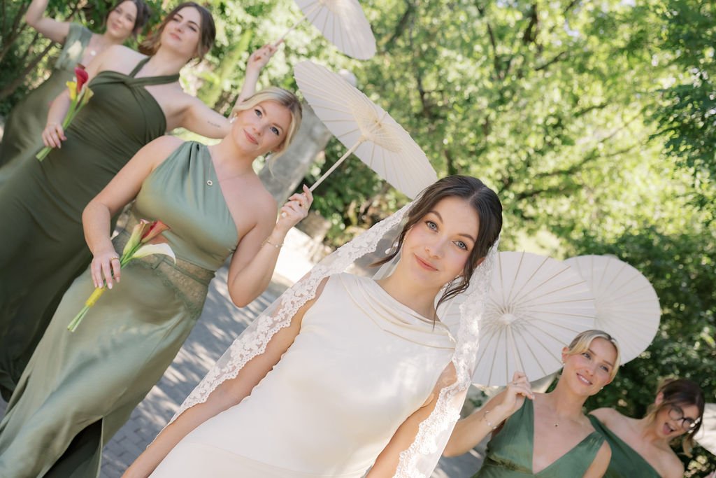 Bride holding a lace parasol with her bridesmaids in green gowns in Provence, natural bridal party makeup by Mathilde Vintze