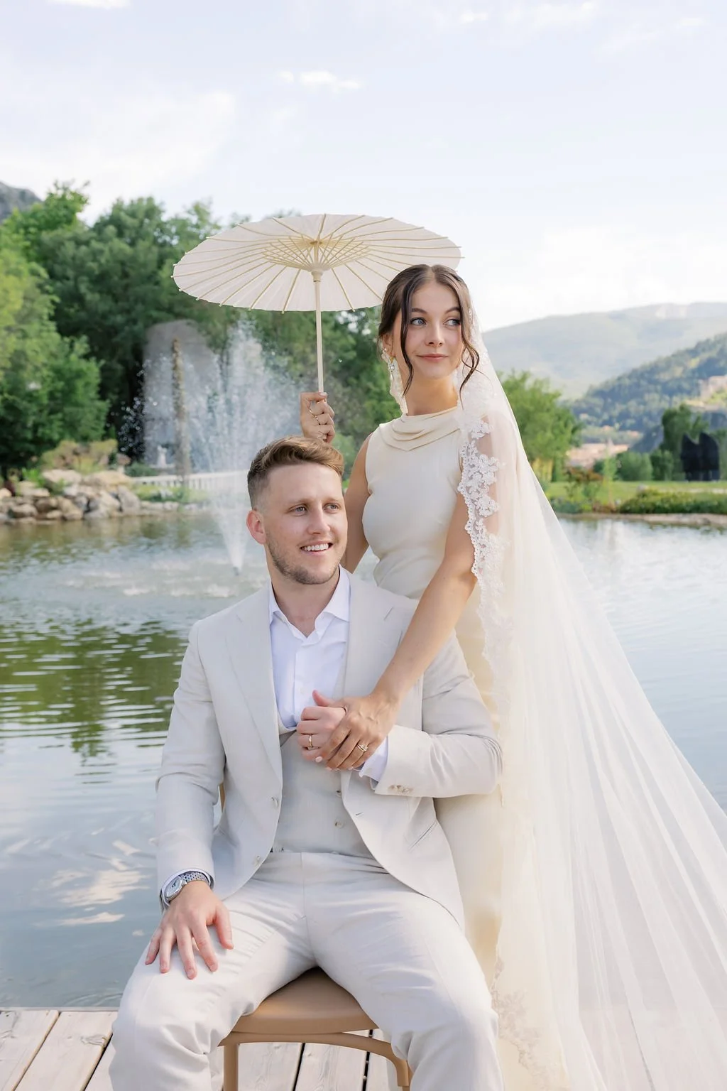 Bride with a lace veil and parasol beside the groom by the water at Mas des Cinq Fontaines, soft natural bridal makeup in Provence by Mathilde Vintze