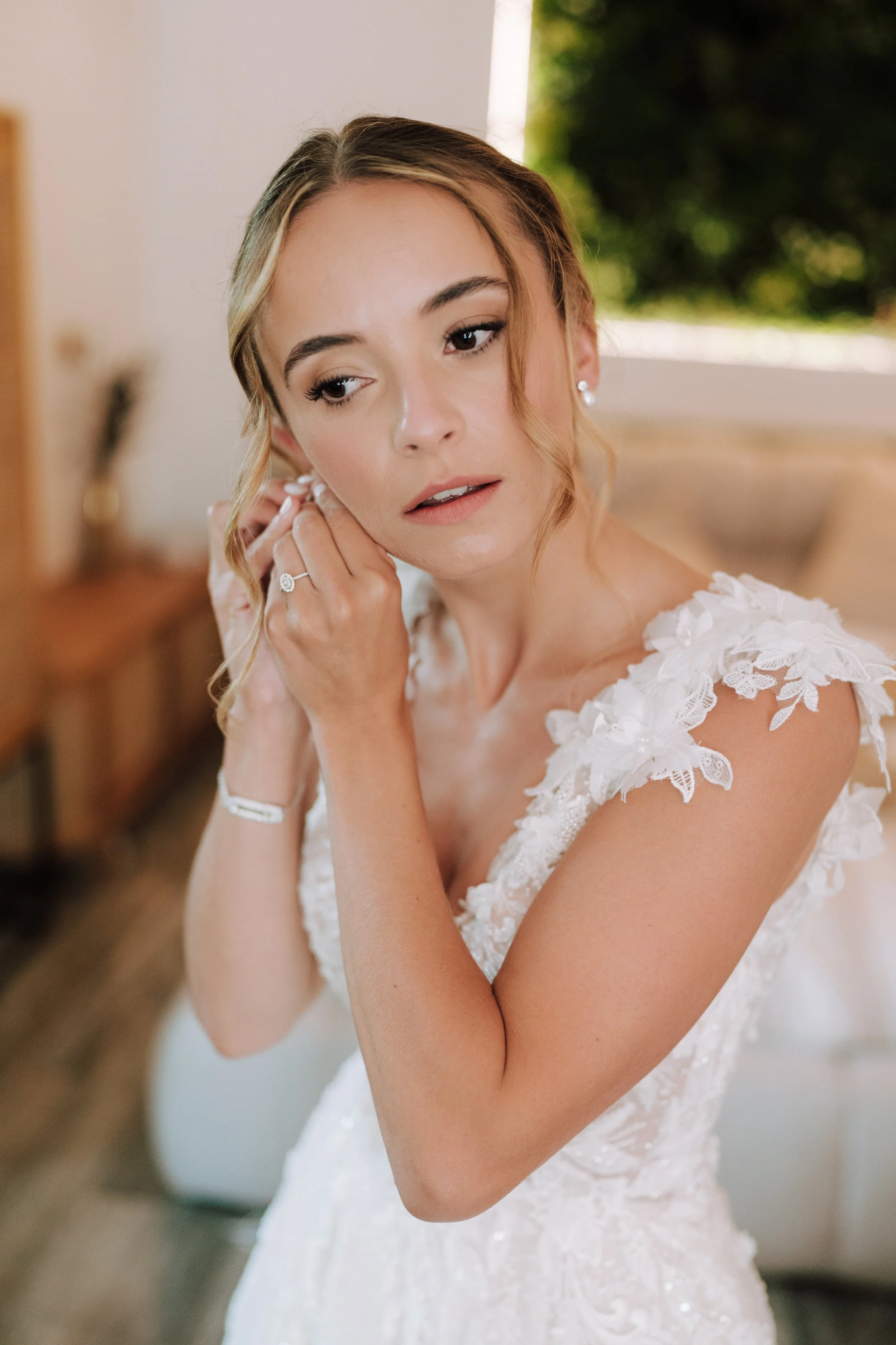 Blonde bride in a lace gown fastening her earring, fresh natural wedding makeup by Mathilde Vintze in Provence
