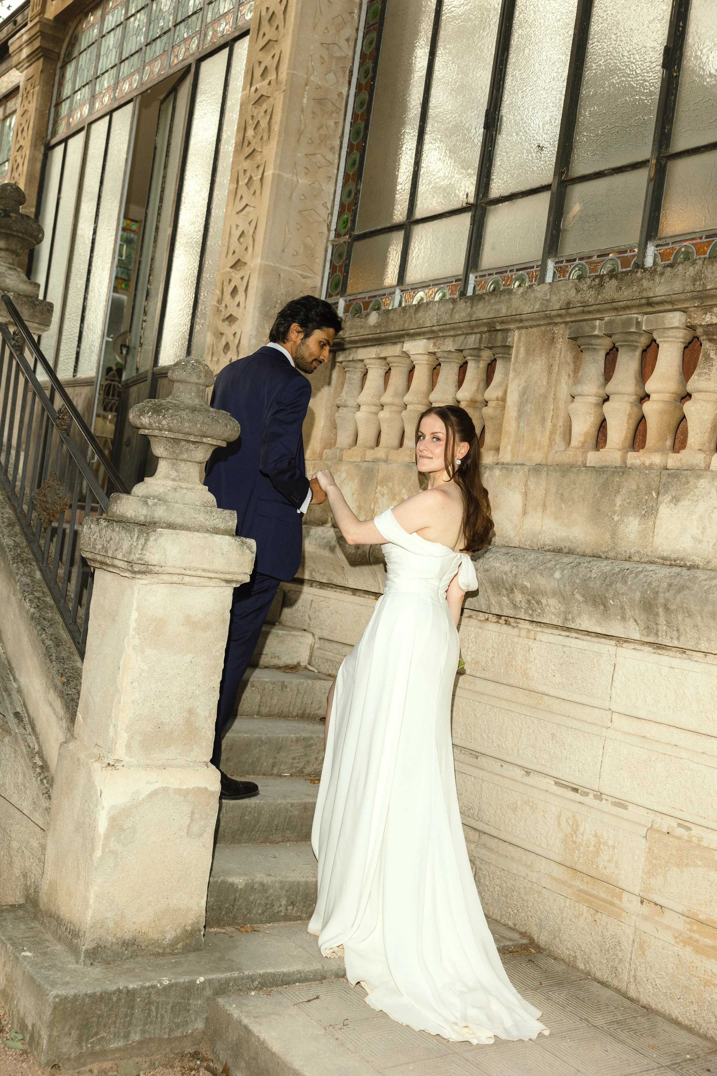 The bride and the groom on a historic stone staircase, the bride in a flowing off-the-shoulder wedding dress with luminous, natural bridal makeup and the groom in a navy suit.