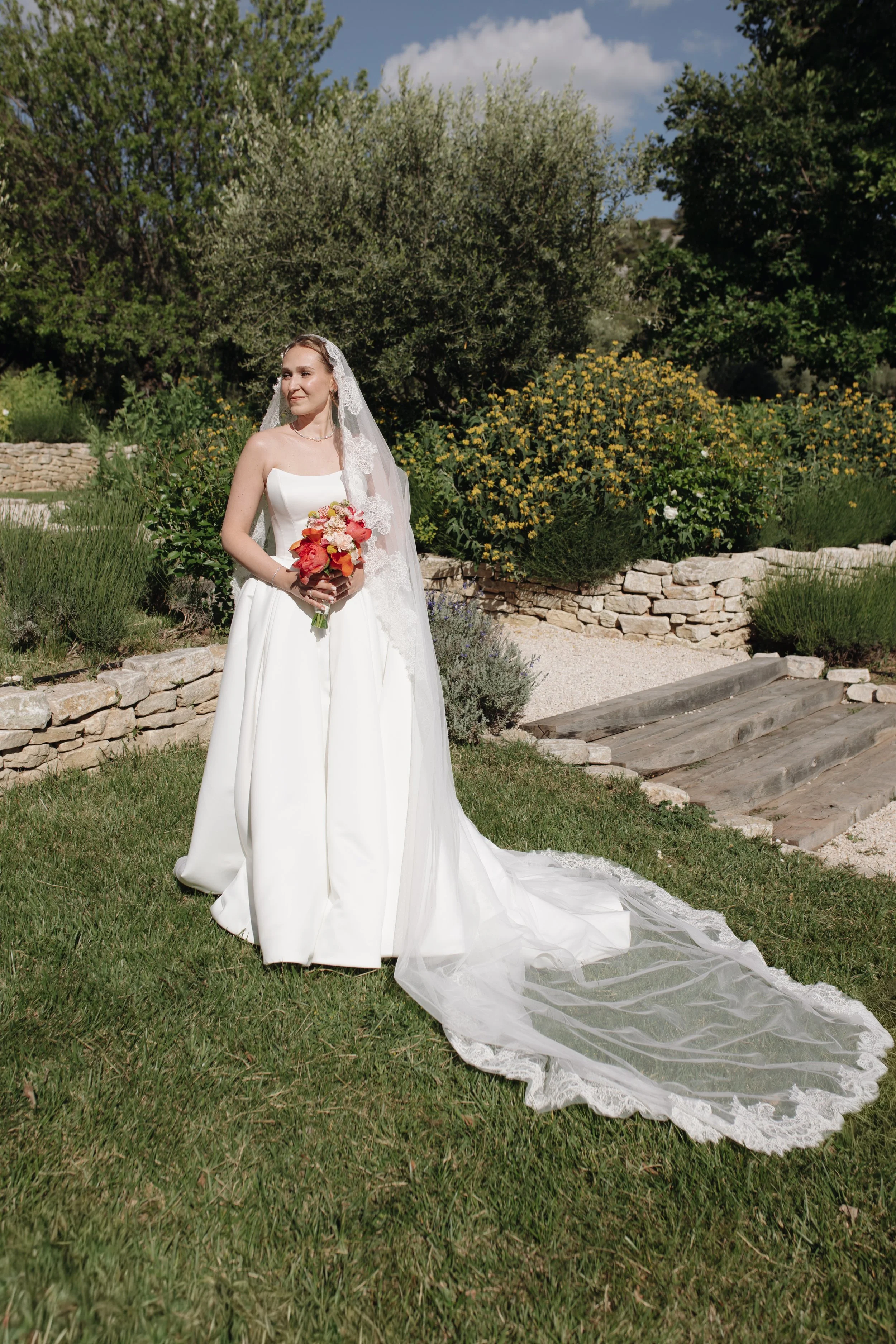 Romantic bride on stone steps with a long-train gown and vibrant bouquet, wearing timeless wedding makeup and loose waves styled by Mathilde Vintze Artistry.