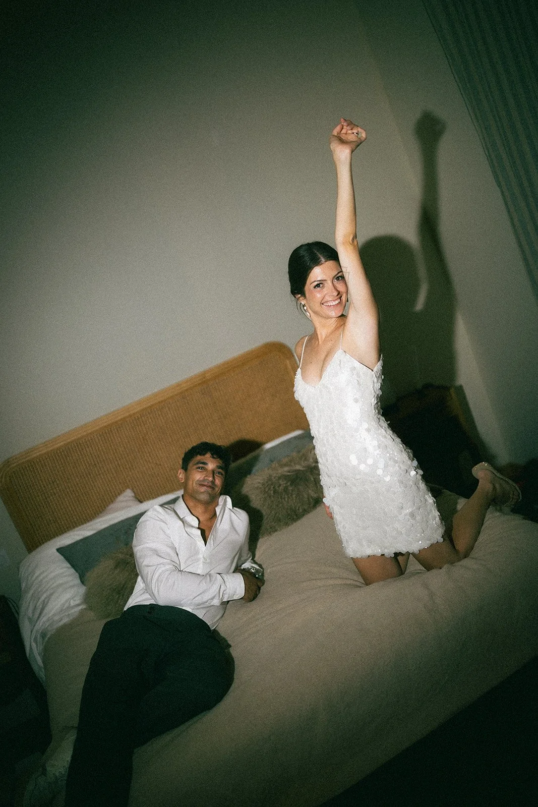The bride dancing on the bed in a short white party dress while the groom laughs, late at night after the celebration. Bridal makeup still intact at the end of the night.