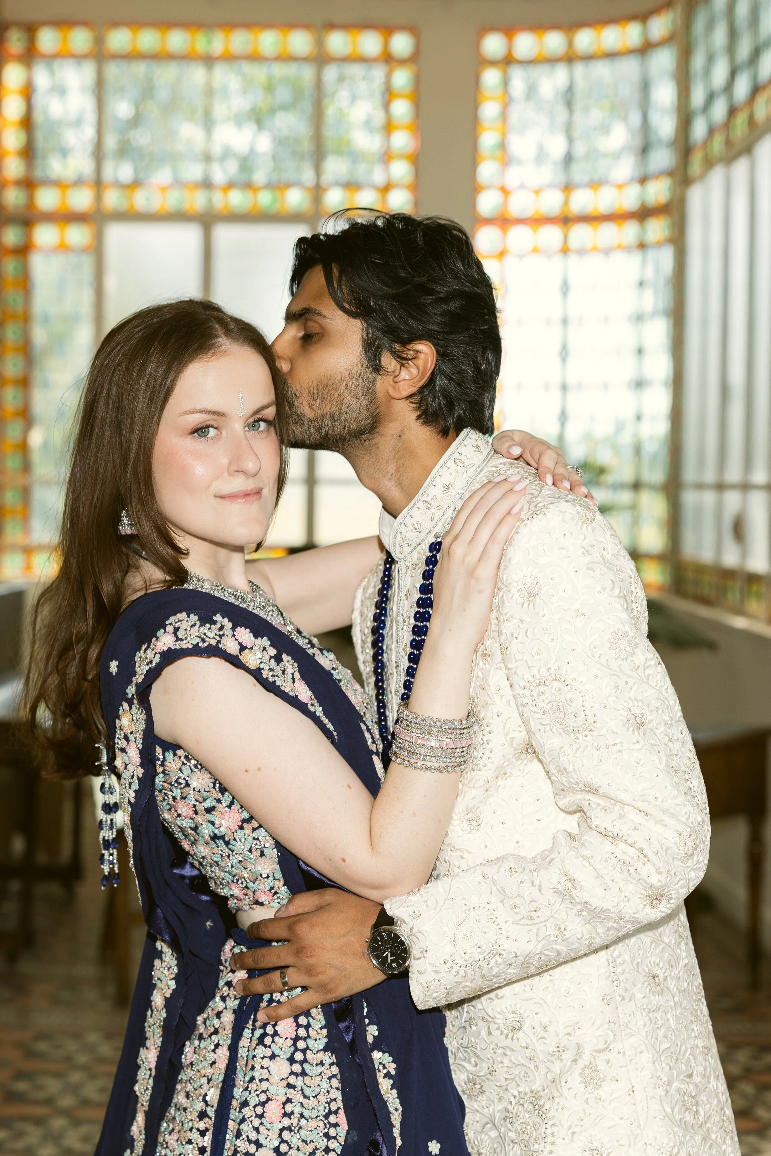 The groom kisses the bride in front of stained-glass windows on their fusion wedding day, the bride in a navy embroidered lehenga and the groom in an ivory sherwani. Natural, glowing Indian bridal makeup by Mathilde Vintze Artistry.