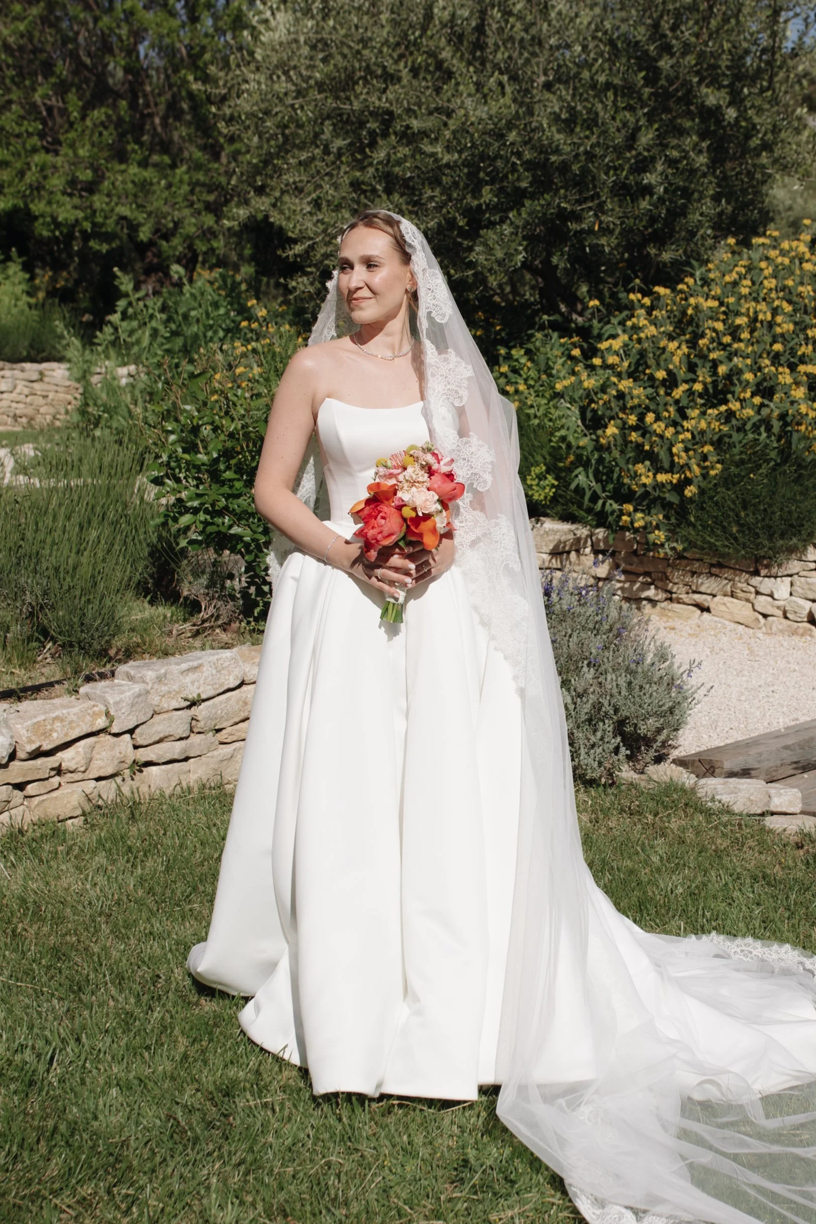 Outdoor bridal portrait featuring soft natural wedding makeup and an elegant updo by Mathilde Vintze Artistry. Romantic countryside bride in a flowing white gown holding a colourful bouquet.