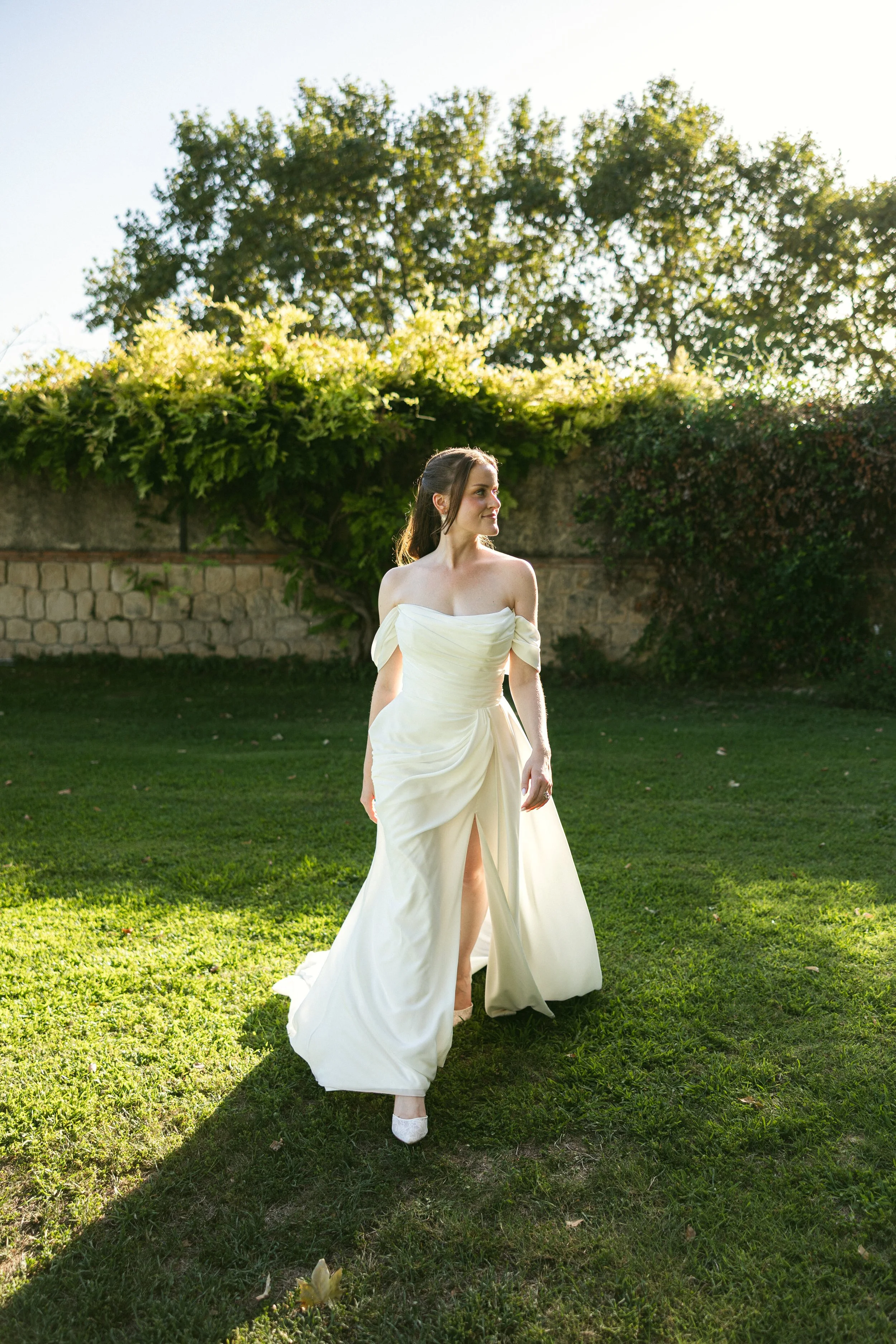 Bride walking across a sunlit garden lawn in a draped off-the-shoulder gown with a leg slit. Fresh, long-lasting bridal makeup made for outdoor weddings in the South of France.