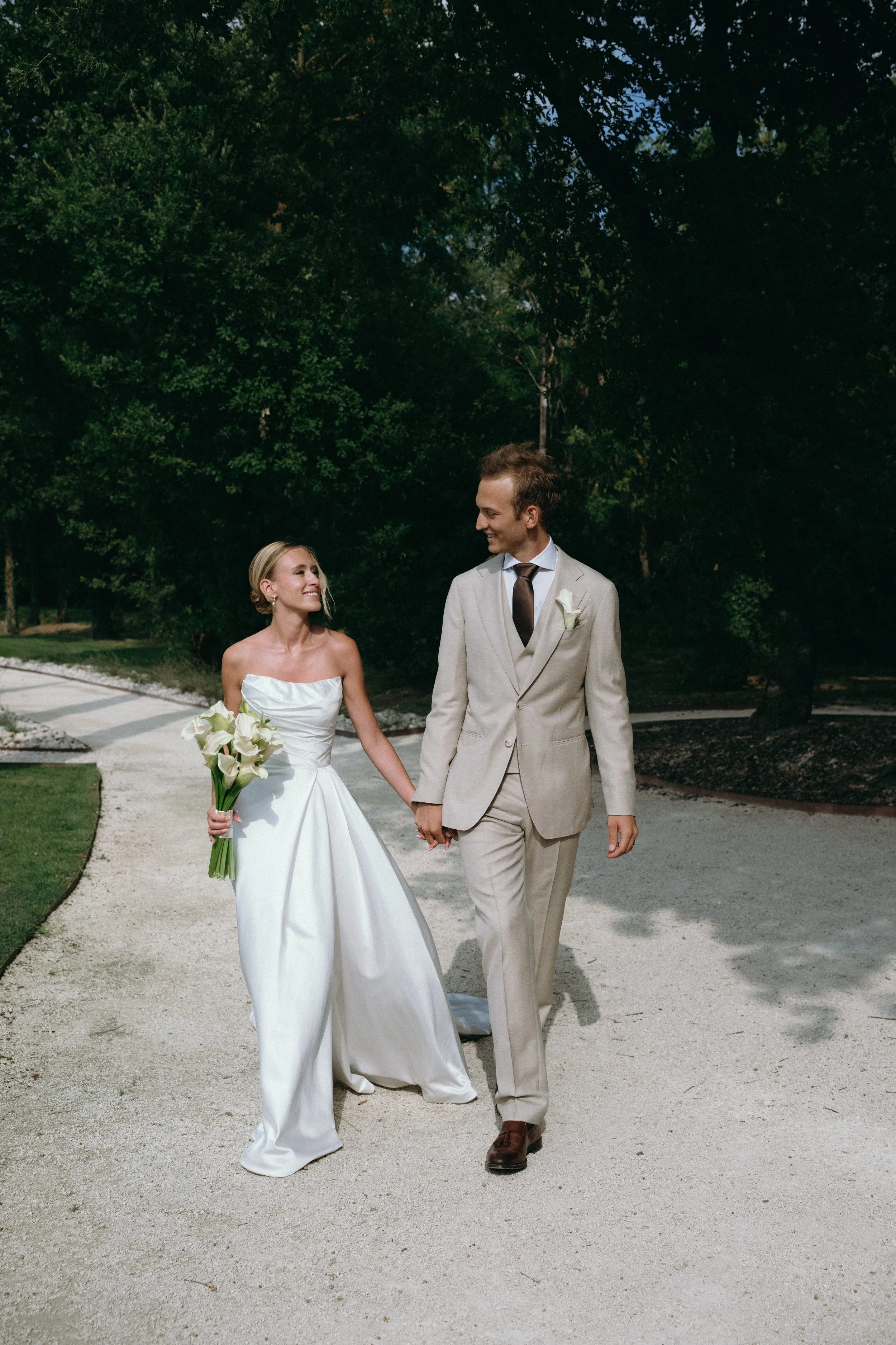 A newlywed couple holding hands and walking outdoors on a gravel path surrounded by trees, with the bride in a strapless white wedding dress holding a bouquet of calla lilies, and the groom in a light-colored suit with a dark tie.