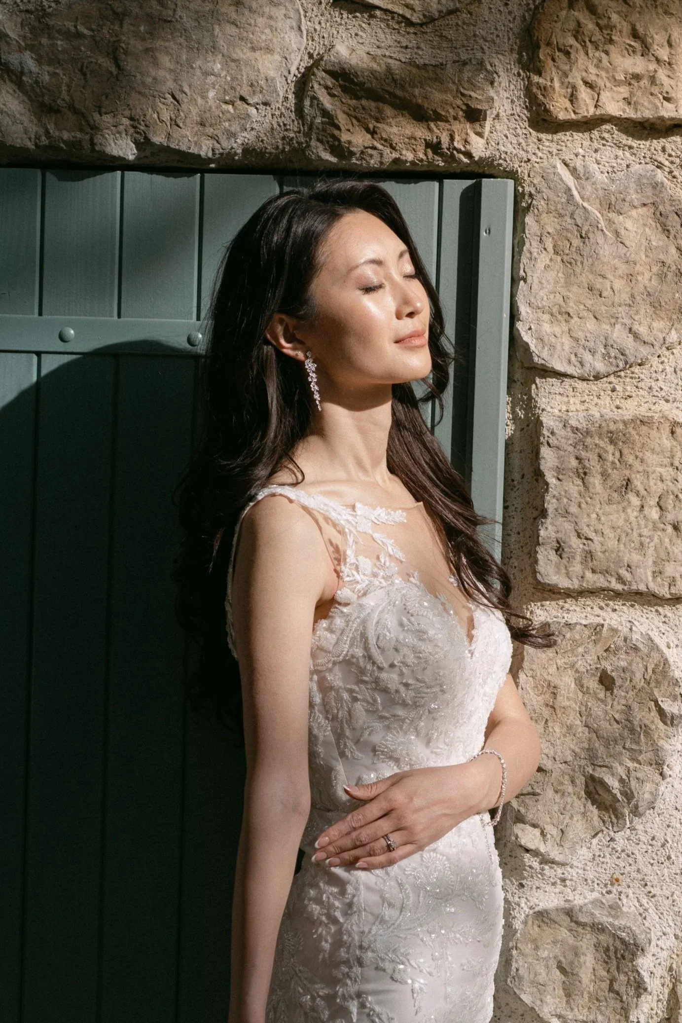 Bride in a lace gown against a stone wall, glowing natural skin and soft bridal makeup in Provence