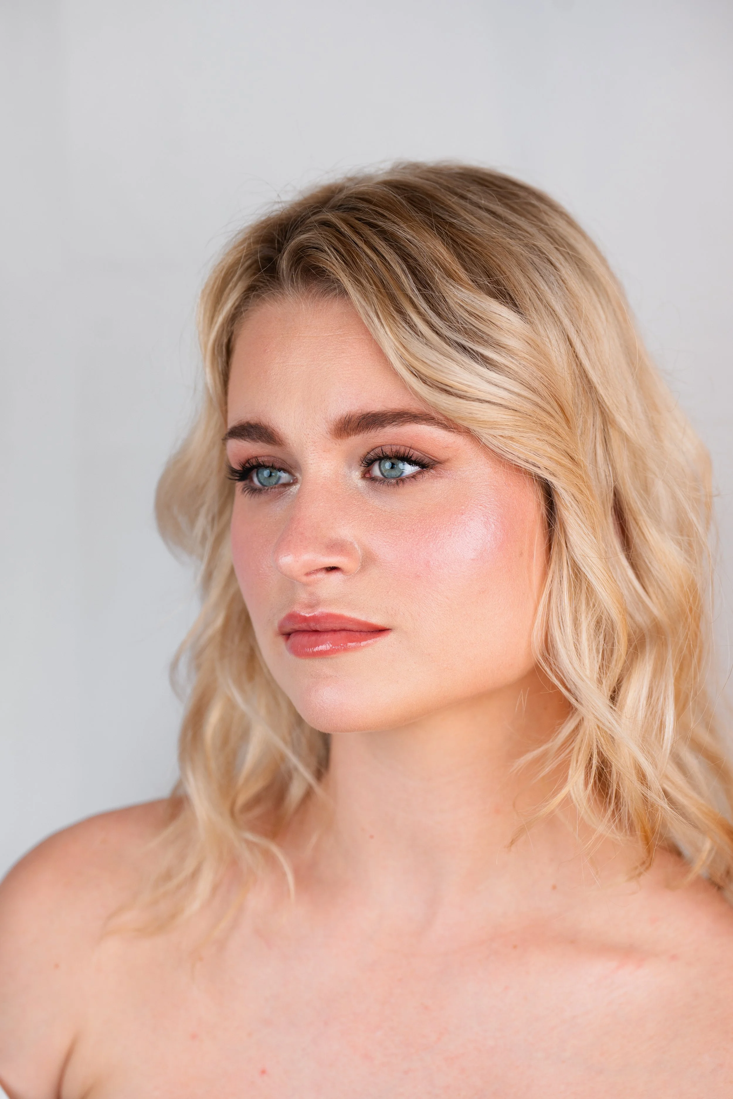 Close-up portrait of a young woman with shoulder-length blonde wavy hair, blue eyes, and natural makeup, looking slightly to the side against a neutral background.