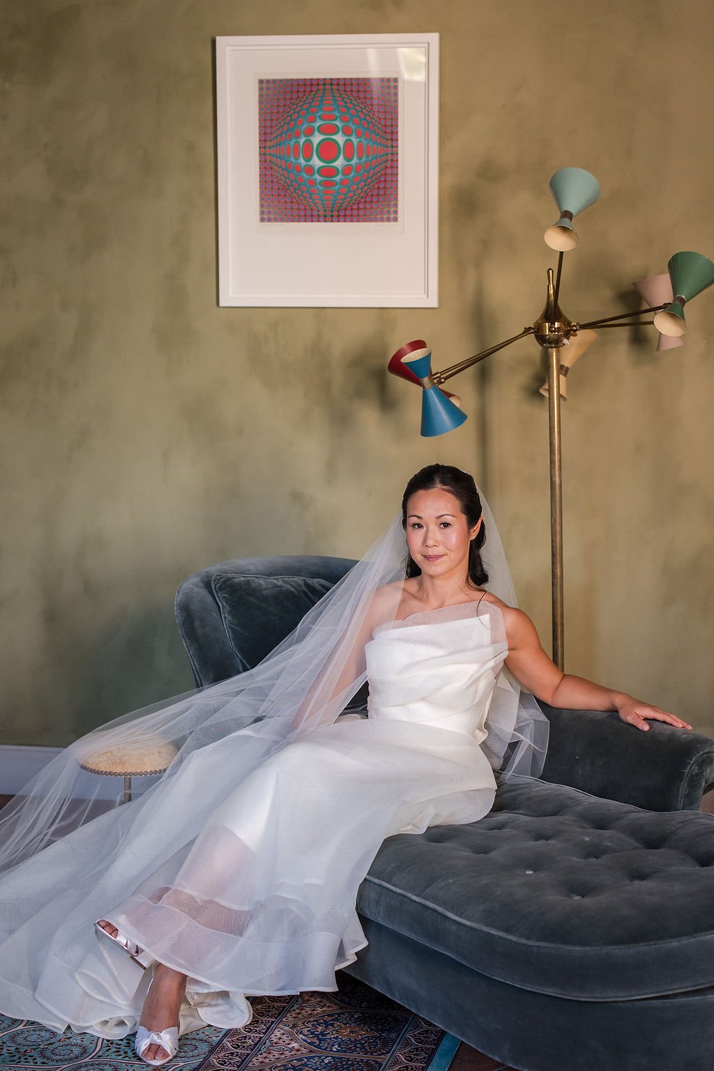 Bride in a one-shoulder wedding gown and long veil on a velvet sofa, elegant natural bridal makeup by Provence makeup artist Mathilde Vintze