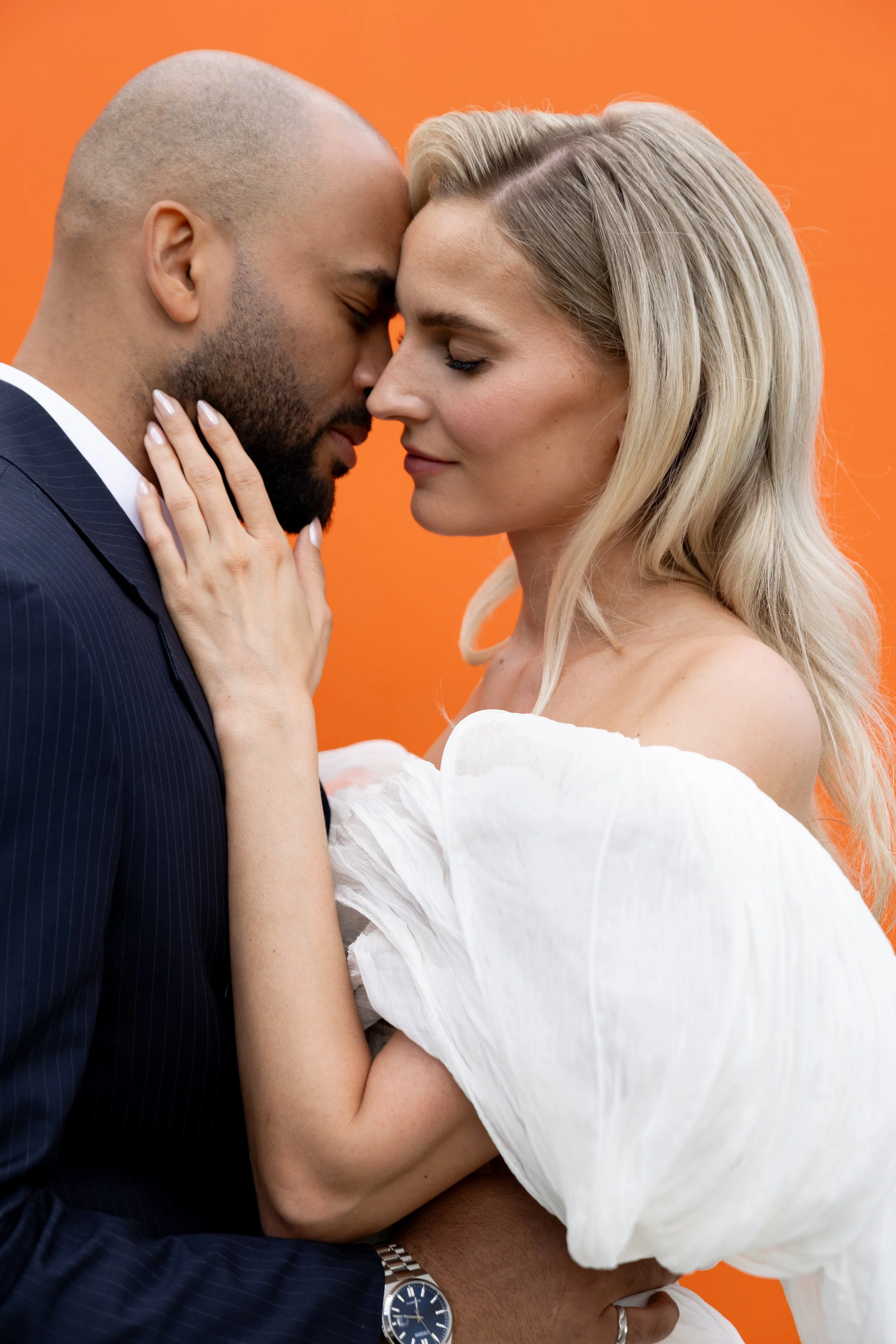Close-up of the bride and groom cheek to cheek against a bright orange wall, radiant long-wearing bridal makeup by Mathilde Vintze