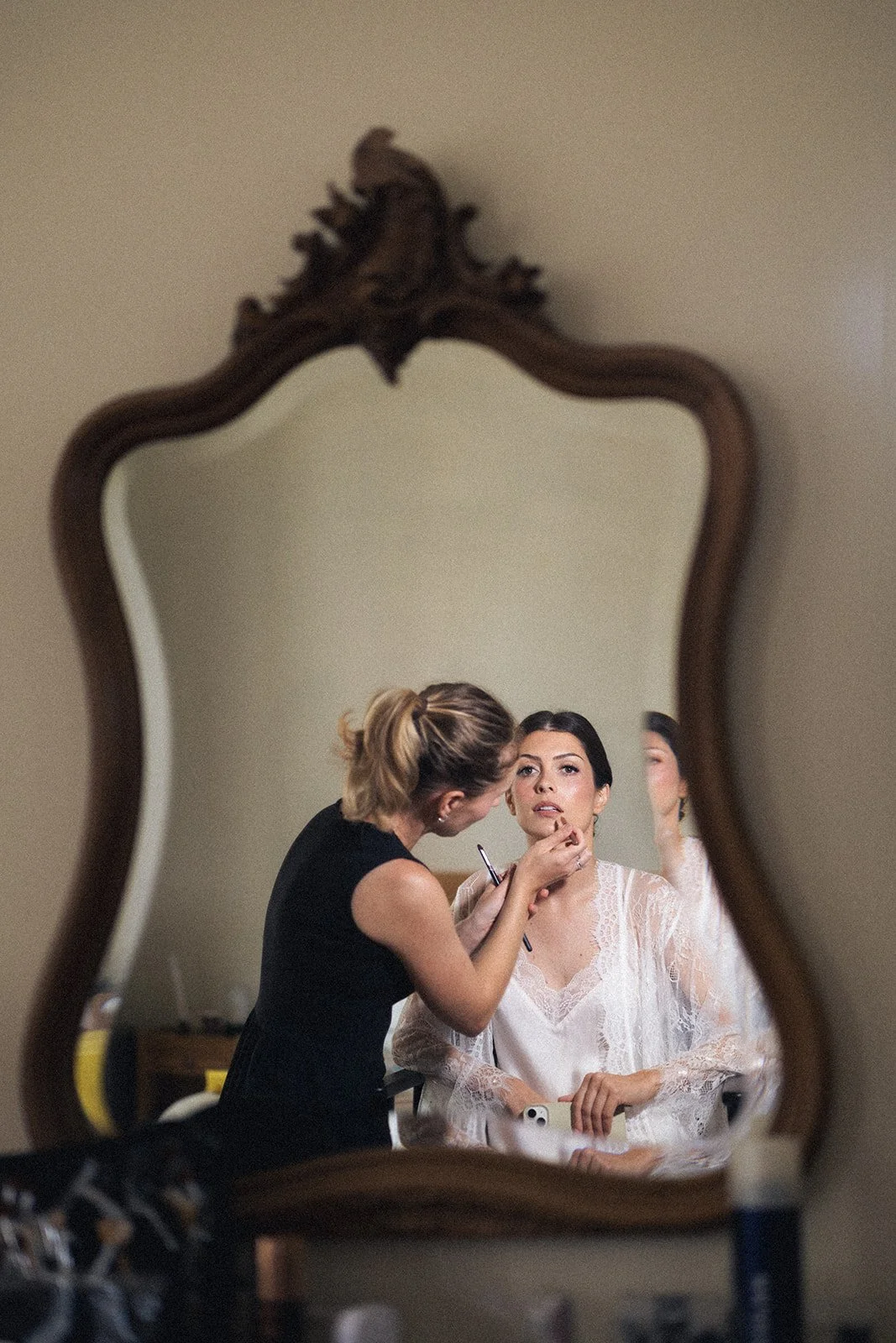 Bridal makeup artist Mathilde Vintze applying the bride's makeup, reflected in an ornate antique mirror during wedding morning preparations in Provence.