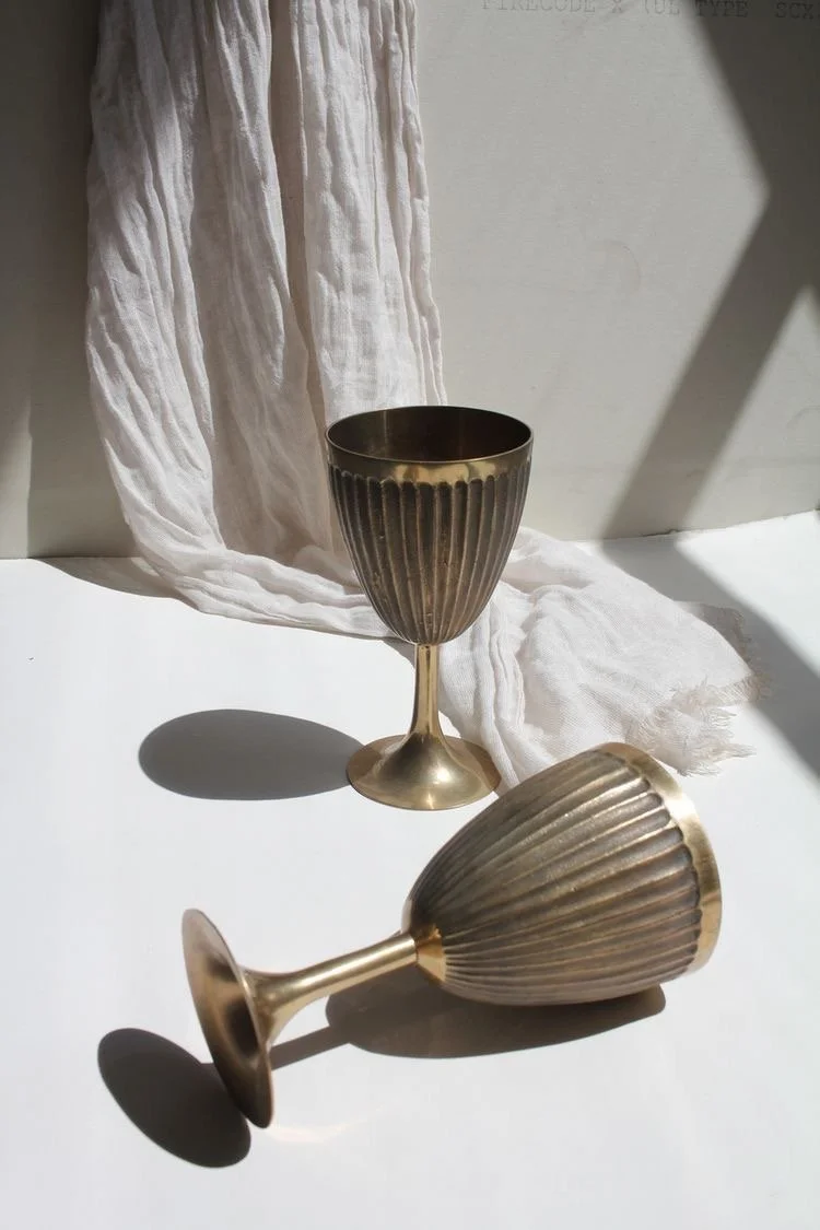 Dos copas doradas de diseño vintage, una de ellas de pie y la otra recostada, sobre superficie blanca con tela blanca y fondo neutro.