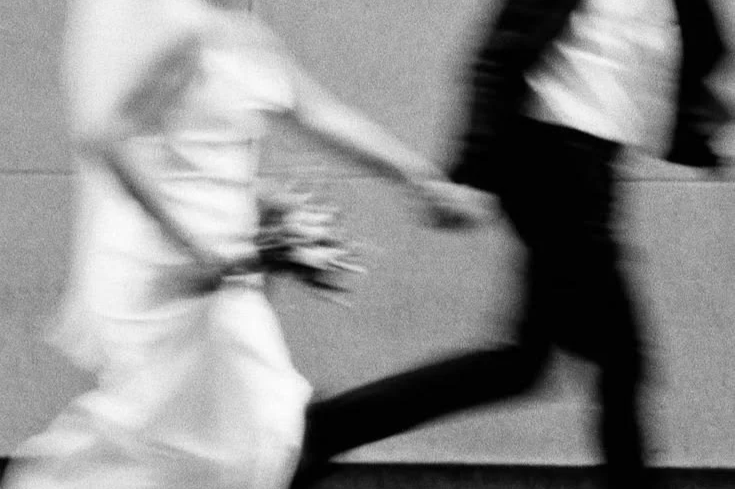 Persona que lleva un vestido blanco y sostiene un ramo, saliendo corriendo con otra persona en un pasillo.