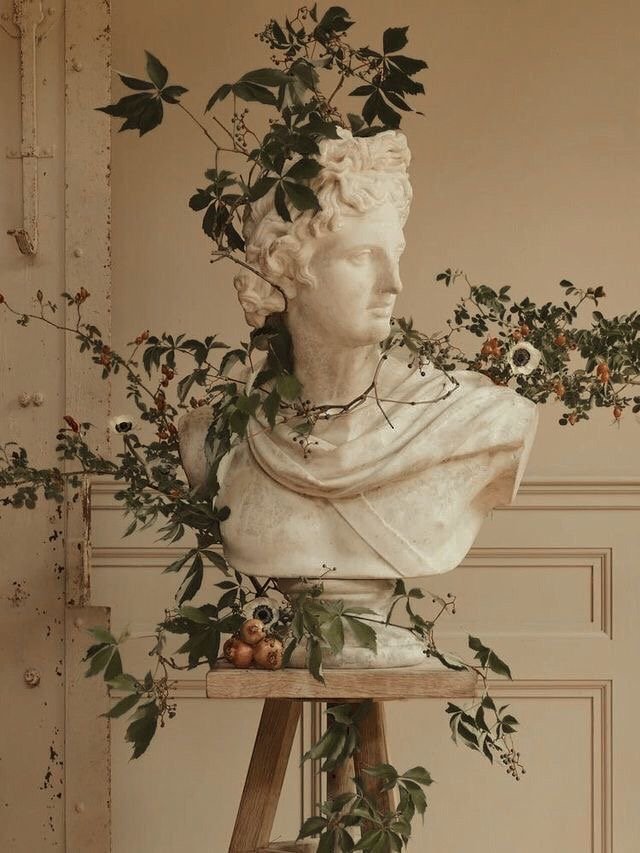 Escultura de piedra de un busto de hombre con cabello rizado, decorada con ramas y hojas verdes, y adornada con pequeñas flores y frutas en la base.