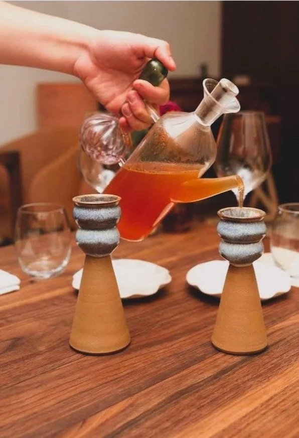 Une personne verse une boisson dans deux tasses sur des supports en forme de cône en terre cuite sur une table en bois.