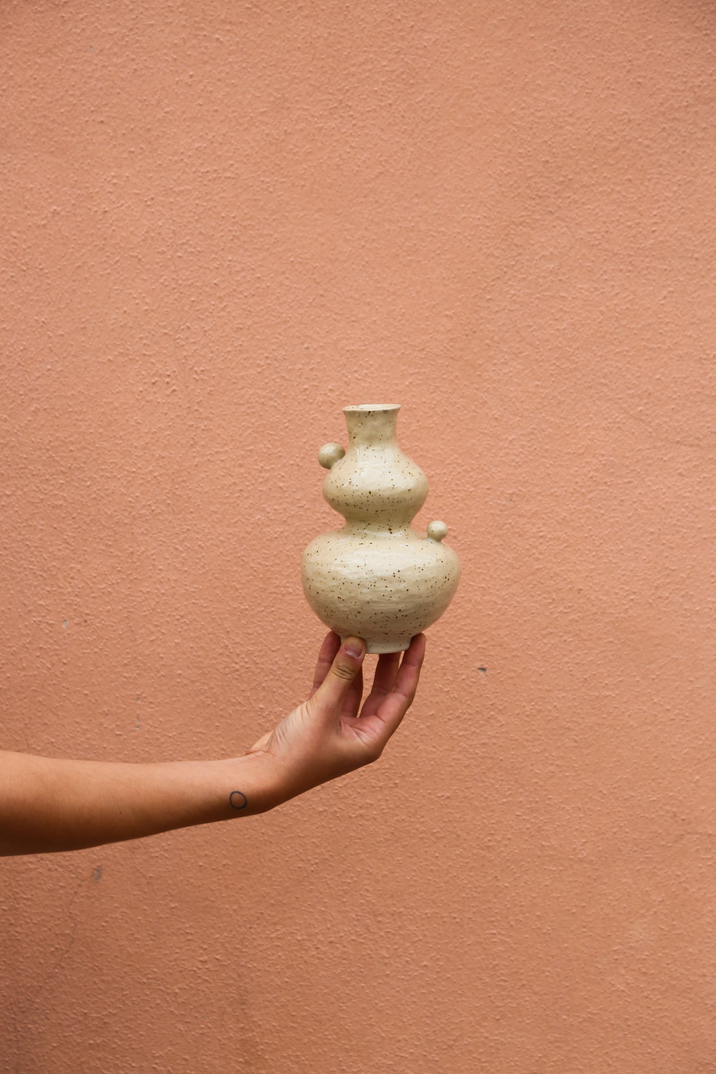 Main tenant une petite statue de céramique beige avec plusieurs formes arrondies et deux petites boules sur le côté, devant un mur rose