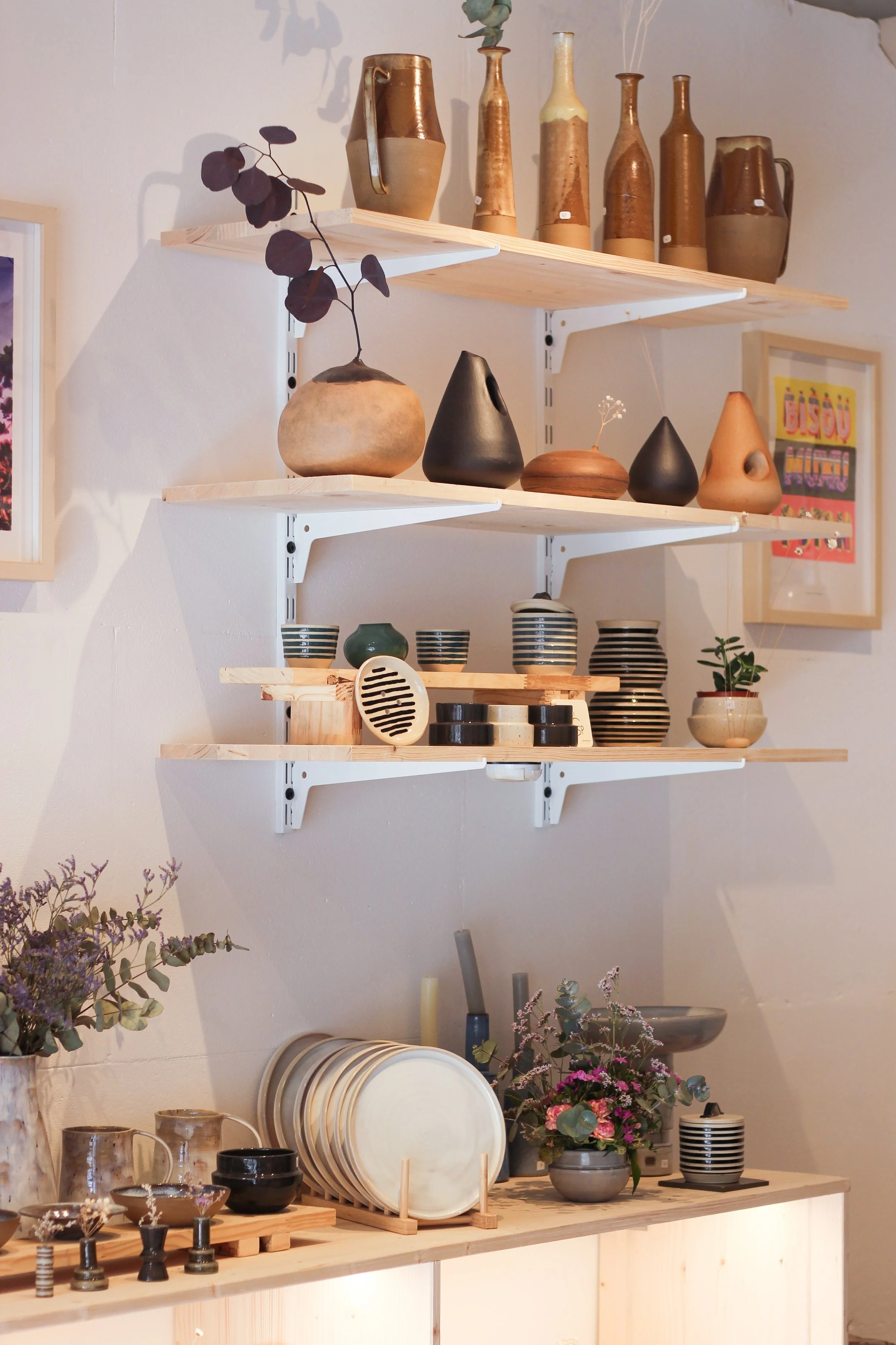 Vases, assiettes et petits objets en céramique exposés sur des étagères en bois fixées à un mur blanc.