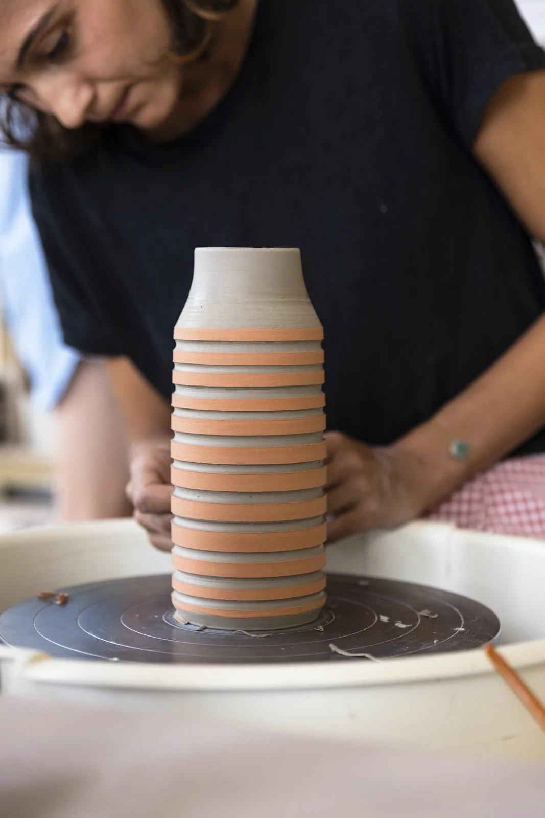 Une femme travaillant sur un tour de poterie, façonnant un vase en argile beige avec des bandes orange autour.