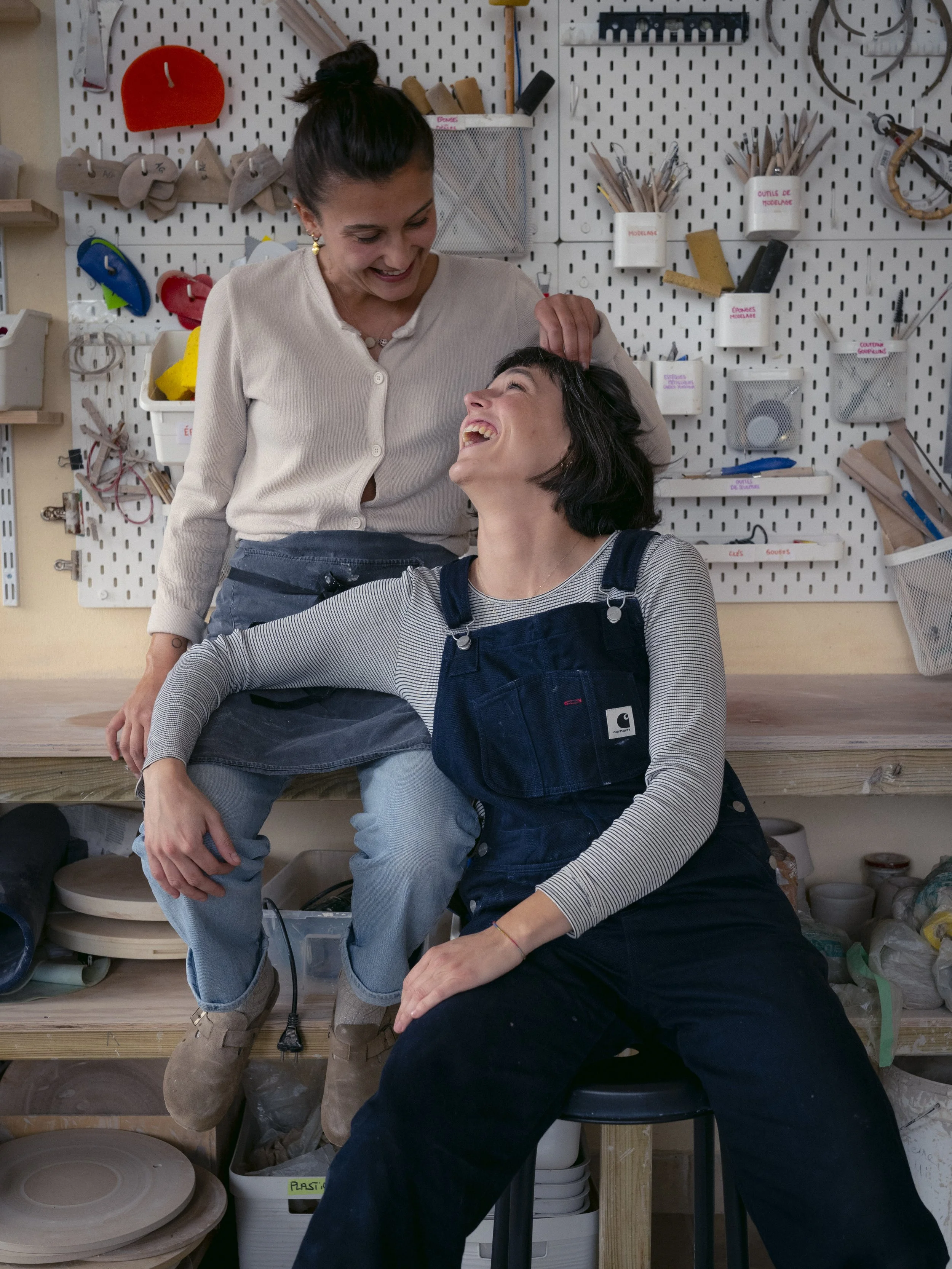 Deux femmes dans un atelier de bricolage, souriantes et en train de rire, l'une debout et l'autre assise sur un tabouret.