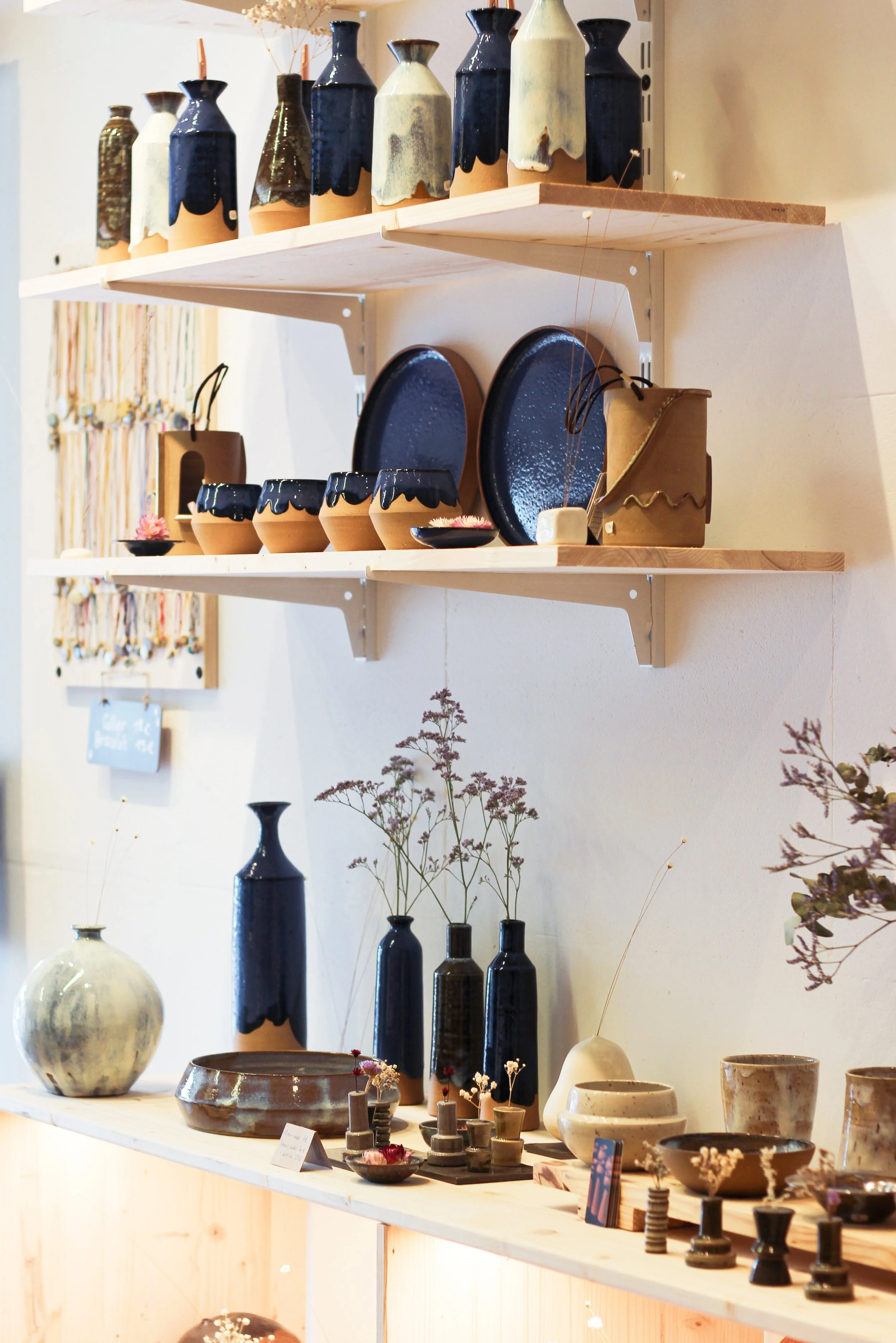 Vases en céramique de différentes formes et couleurs, plates en coquillage, éléments décoratifs en céramique, fleurs séchées dans certains vases, disposés sur des étagères en bois dans une boutique d'artisanat.