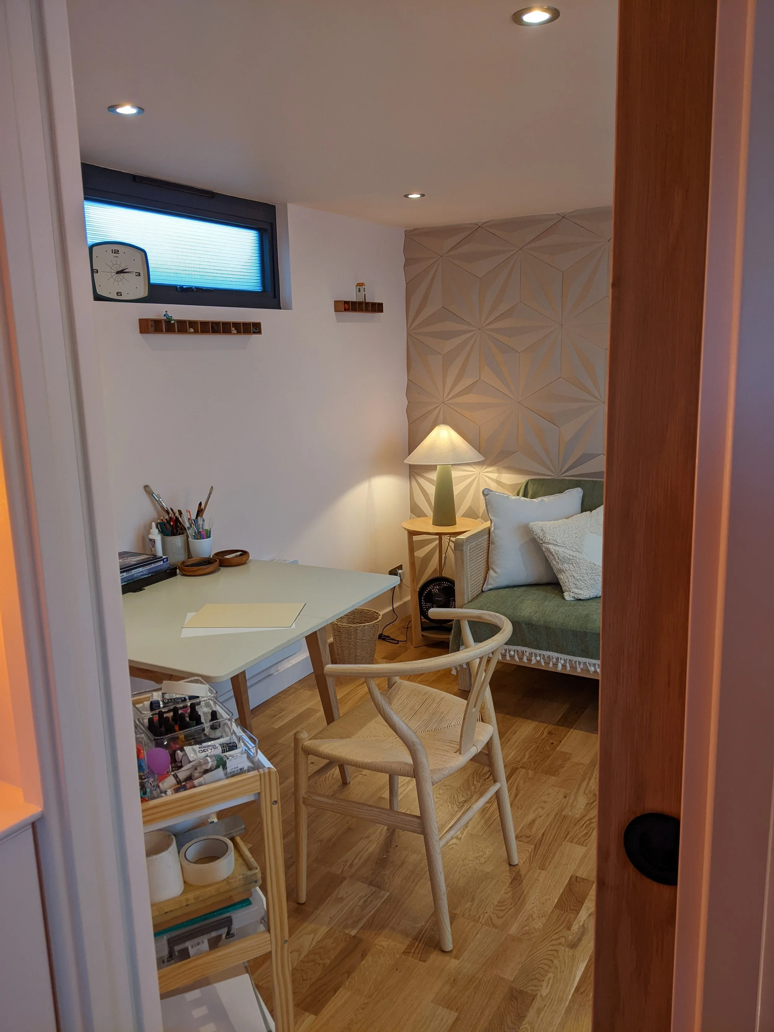 A cozy room with a small window, a white desk with art supplies, a wooden chair, a green sofa with pillows, a lamp, and decorative wall panels.
