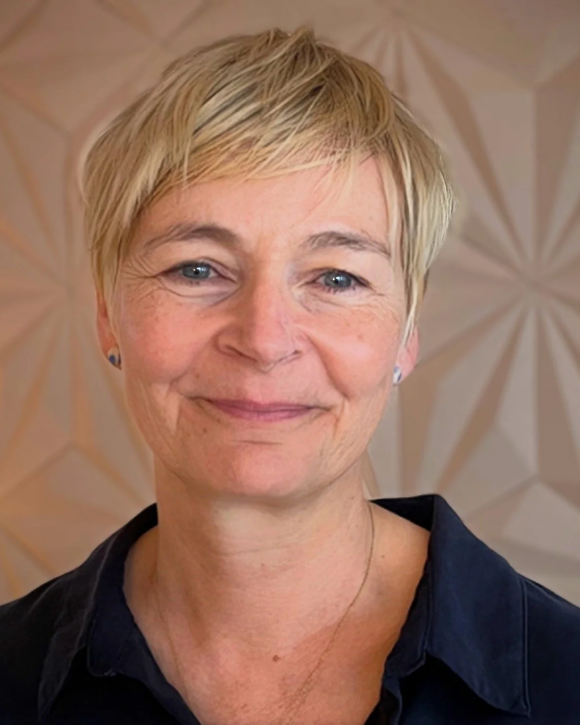 A smiling middle-aged woman with short blonde hair, wearing a dark collared shirt, standing in front of a geometric patterned background.