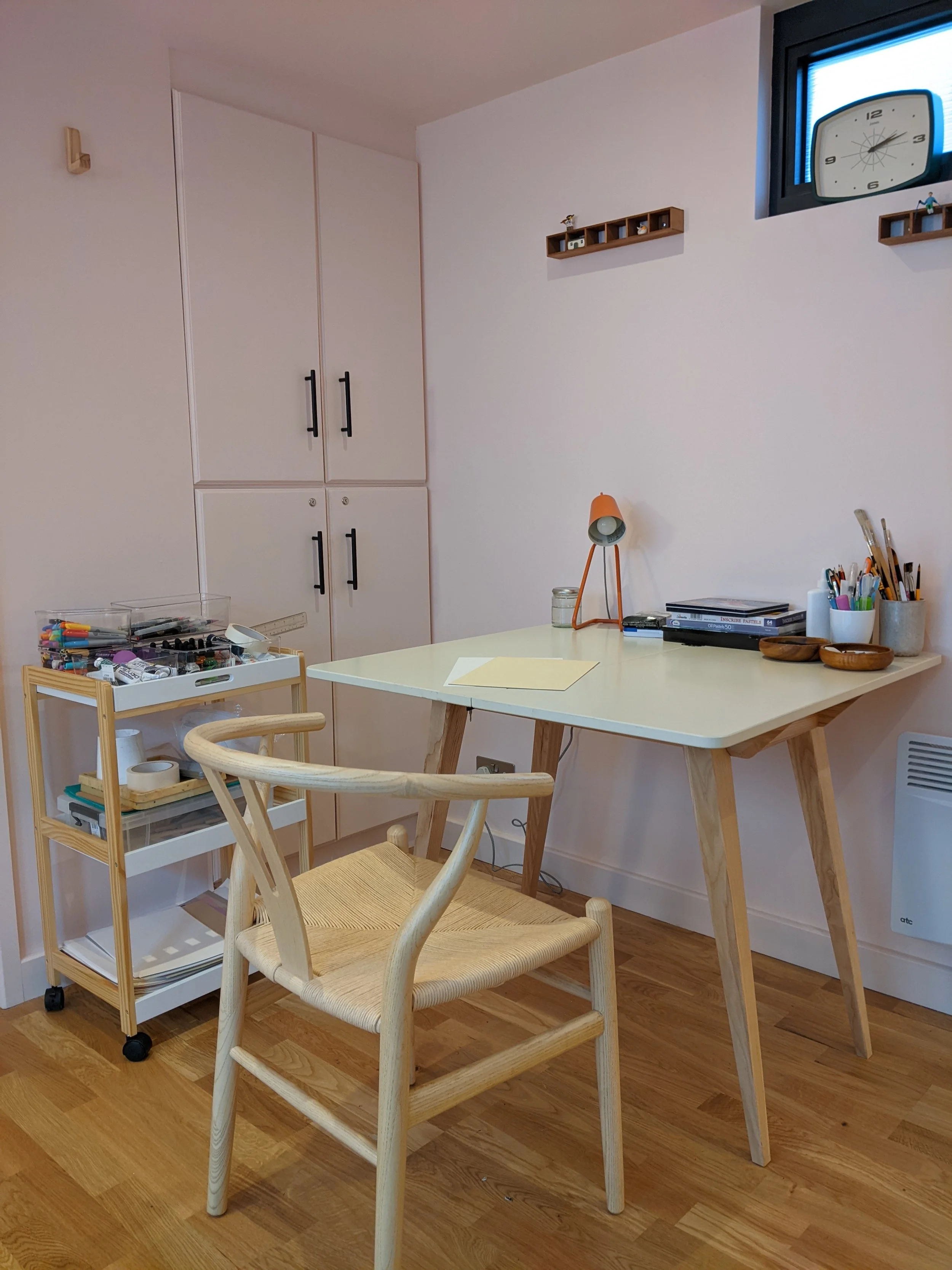A home office or study corner with a wooden desk, a wooden chair with woven seat, a small cart with supplies, a wall-mounted wooden shelf, and a window with a clock.